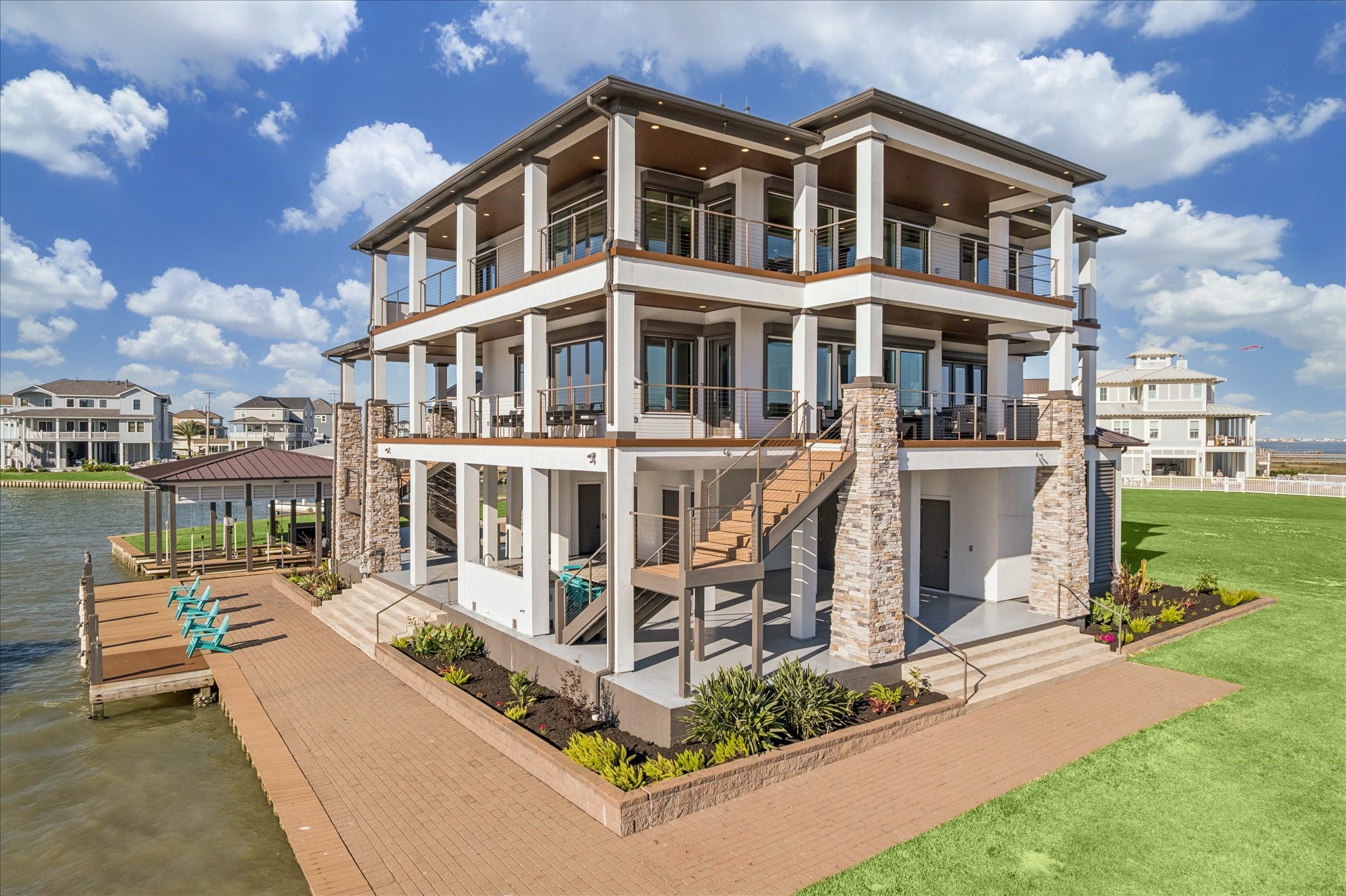 A premier outdoor retreat, featuring expansive paver pathways and manicured landscaping that frame a large shaded dockside patio. Above, multi-level covered balconies accented by stone columns and sleek cable railings offer architectural elegance and panoramic views. The seamless flow from the outdoor spaces creates the ultimate waterfront setting for entertaining