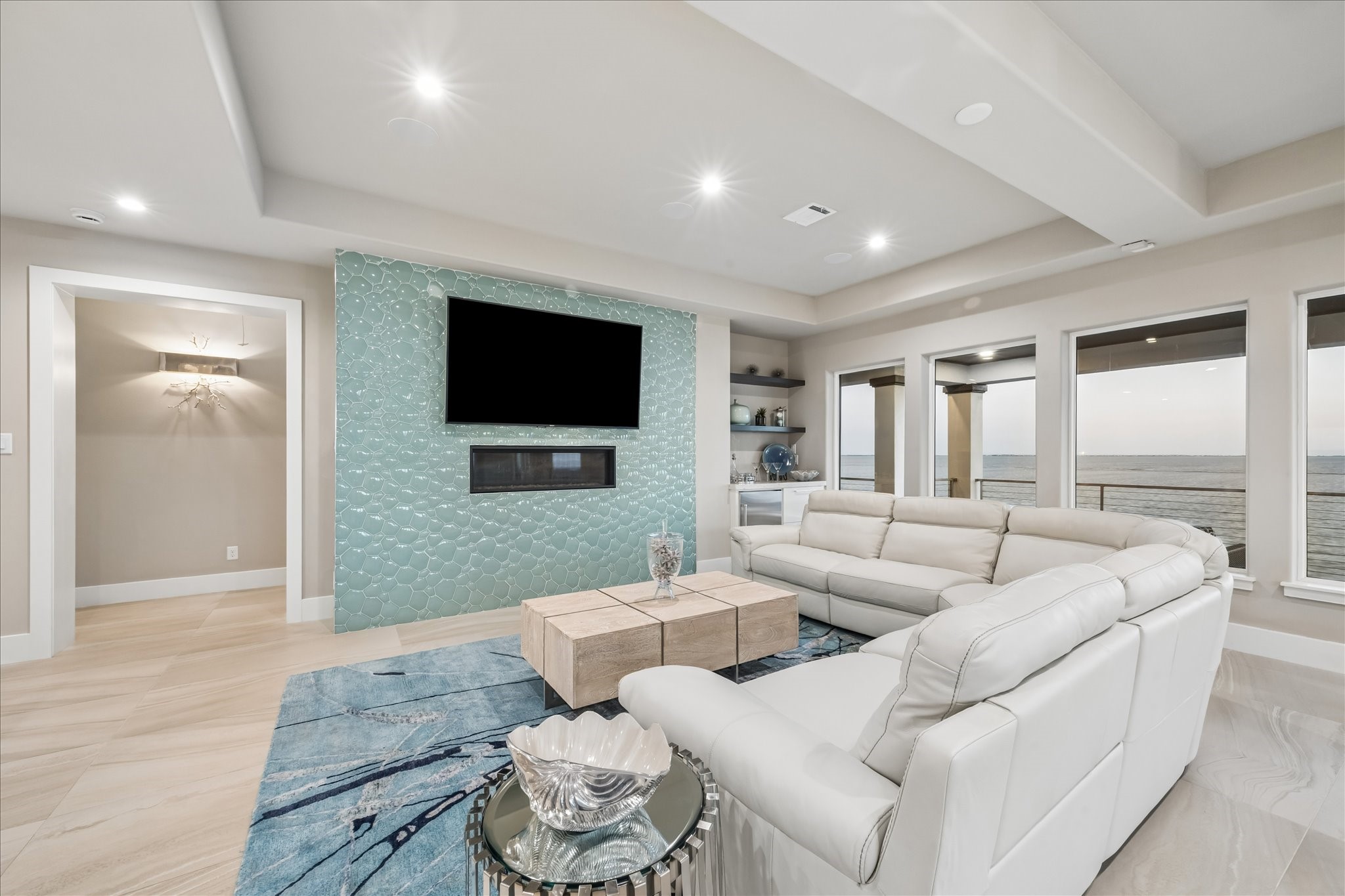 Architectural details throughout the space elevate its modern coastal design, featuring recessed tray ceilings with integrated lighting, a built-in dry bar, and a mosaic-tiled accent wall with a linear fireplace