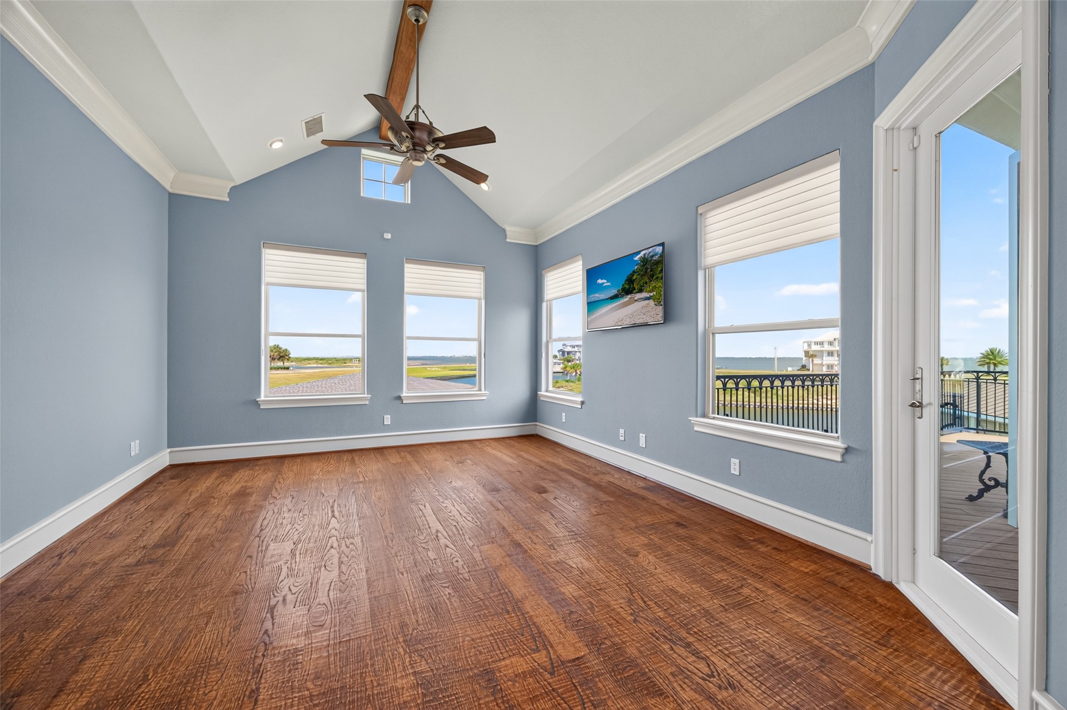 Spacious room with hardwood floors and light blue walls, featuring large windows offering stunning water views and abundant natural light, complemented by a stylish ceiling fan and access to a balcony.