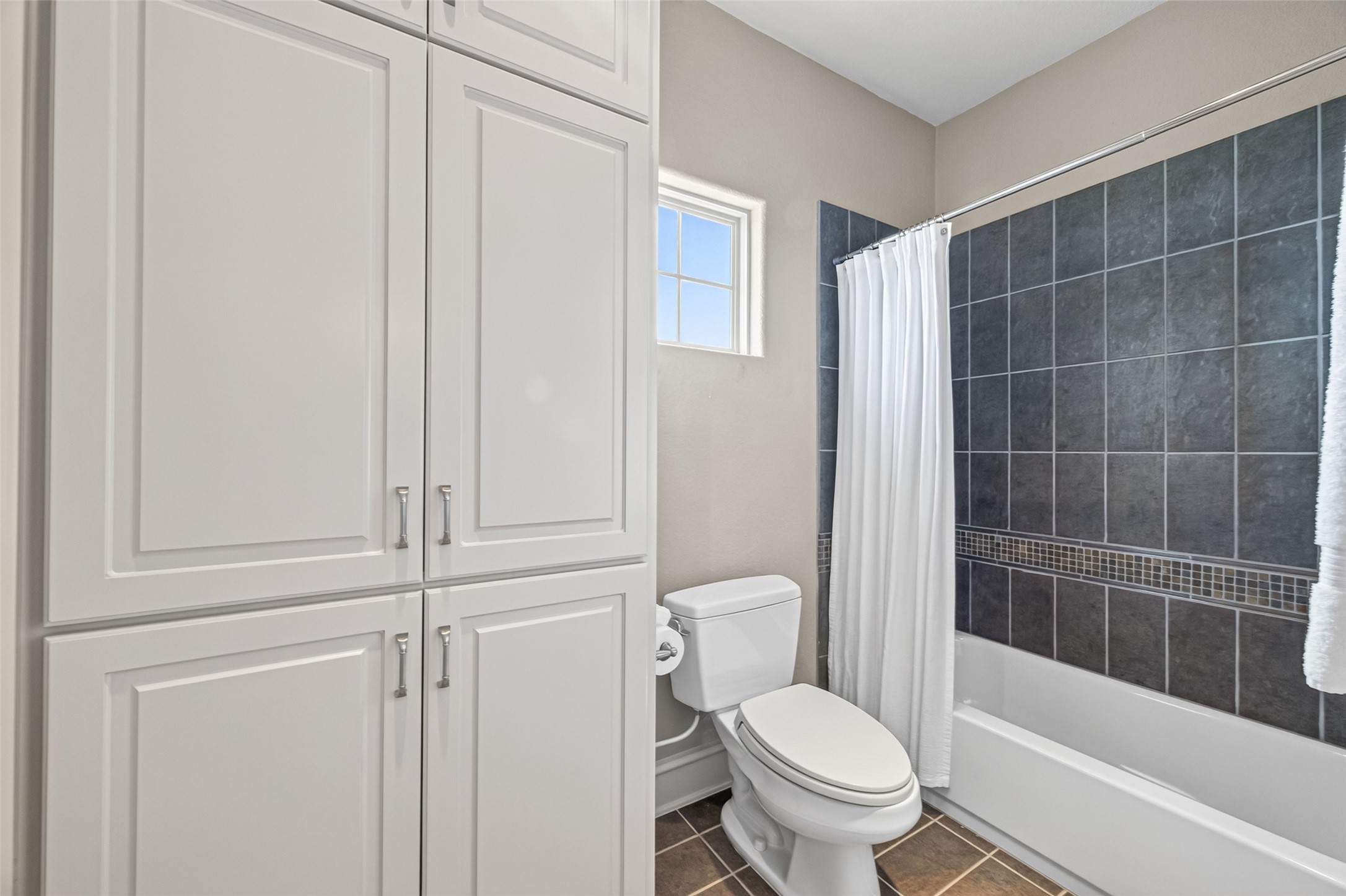Bright bathroom featuring elegant dark tile in the shower, abundant cabinet storage, and a convenient window for natural light.