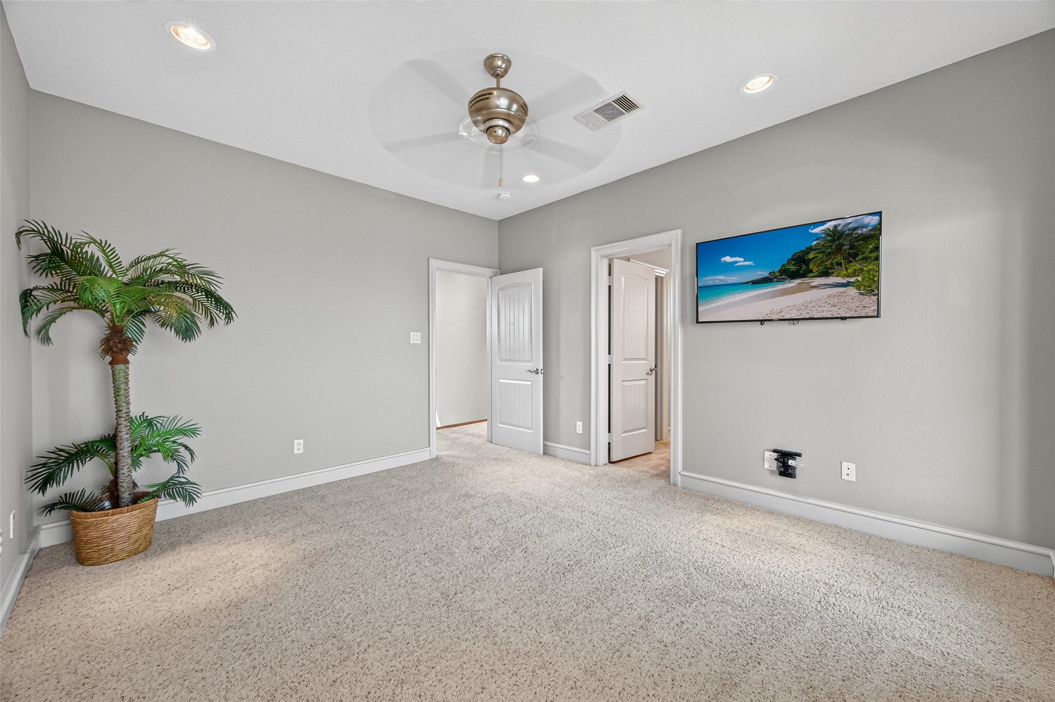 Spacious bedroom featuring plush carpeting, a modern ceiling fan, and a wall-mounted TV, with soft neutral walls and abundant natural light.