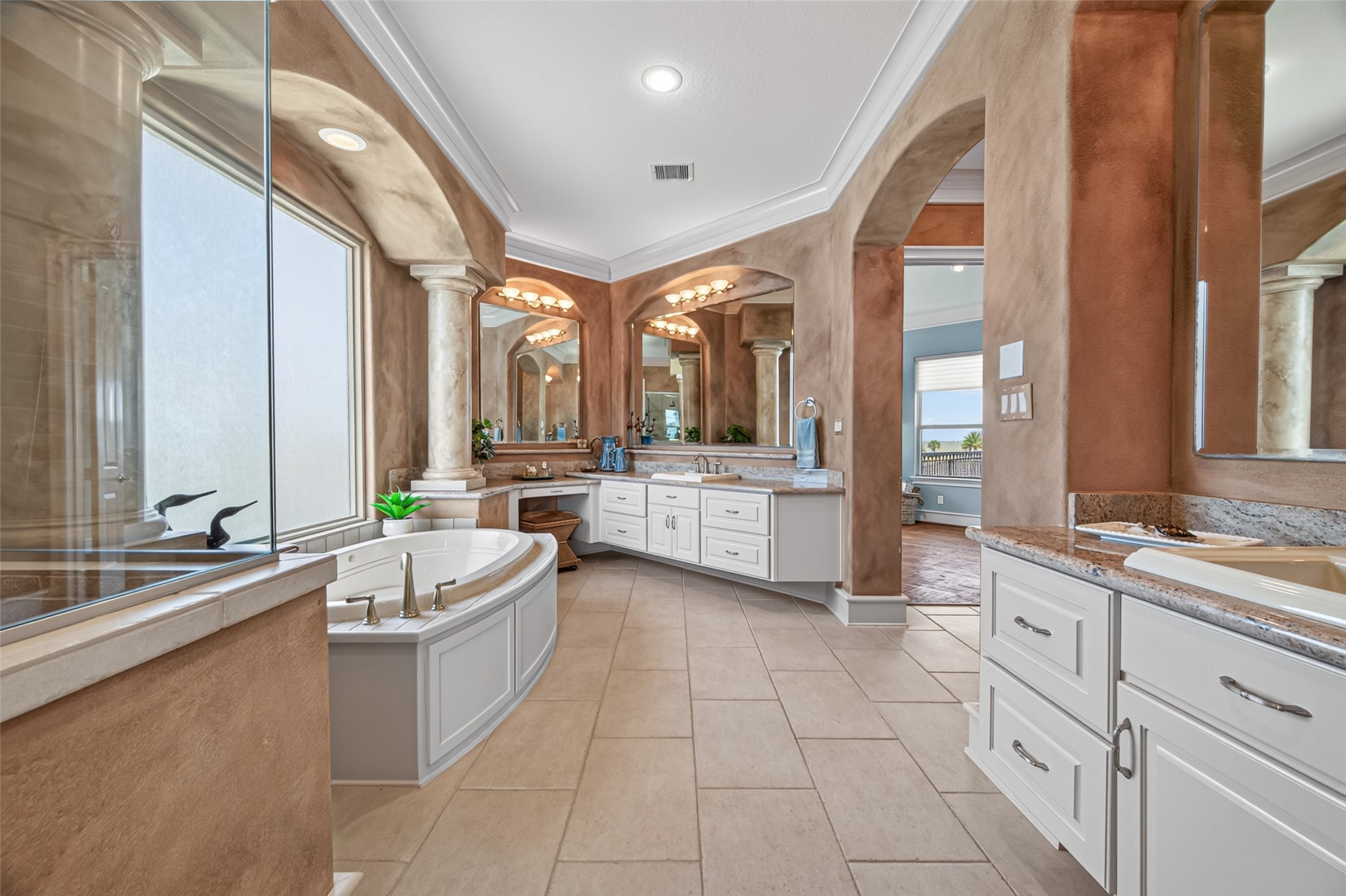 Luxurious bathroom featuring a spacious soaking tub, elegant columns, dual vanities with granite countertops, and a soothing ambiance with warm tones and natural light.