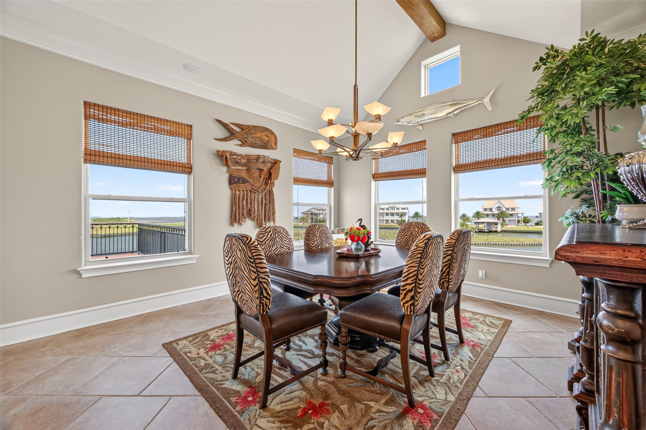 Elegant dining room with high ceilings, natural light, and a chic chandelier. Features stylish zebra-patterned chairs, decorative wall art, and expansive windows offering scenic water views.