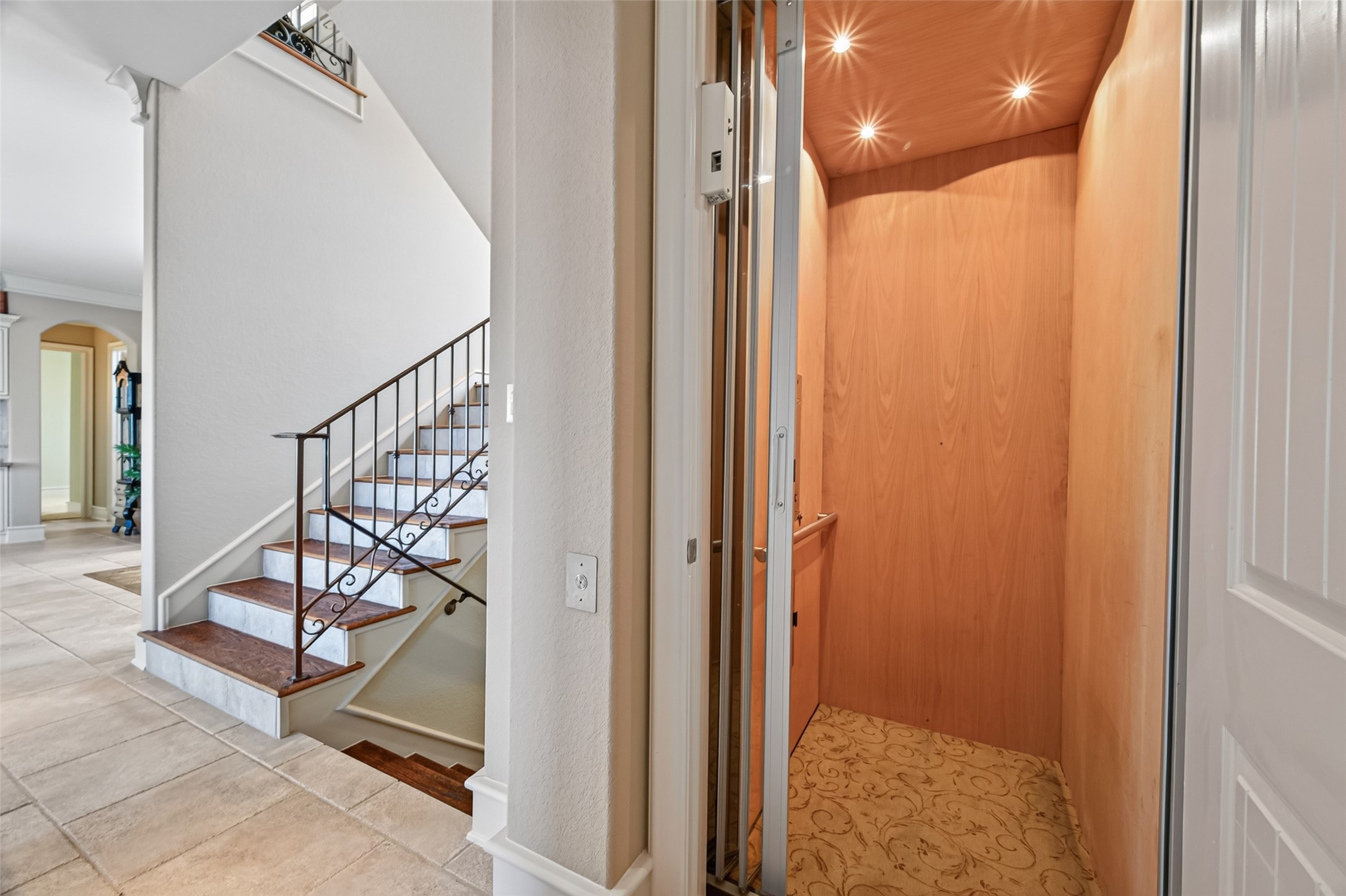 Elegant home featuring a convenient in-house elevator next to a stylish staircase with wrought iron railing, set against a backdrop of light tile flooring and open archways.