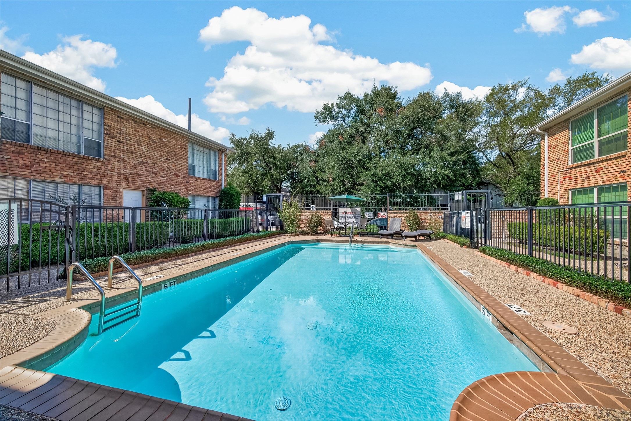 Dive into relaxation with this inviting community pool, surrounded by lush greenery and charming brick buildings, perfect for unwinding on sunny days.
