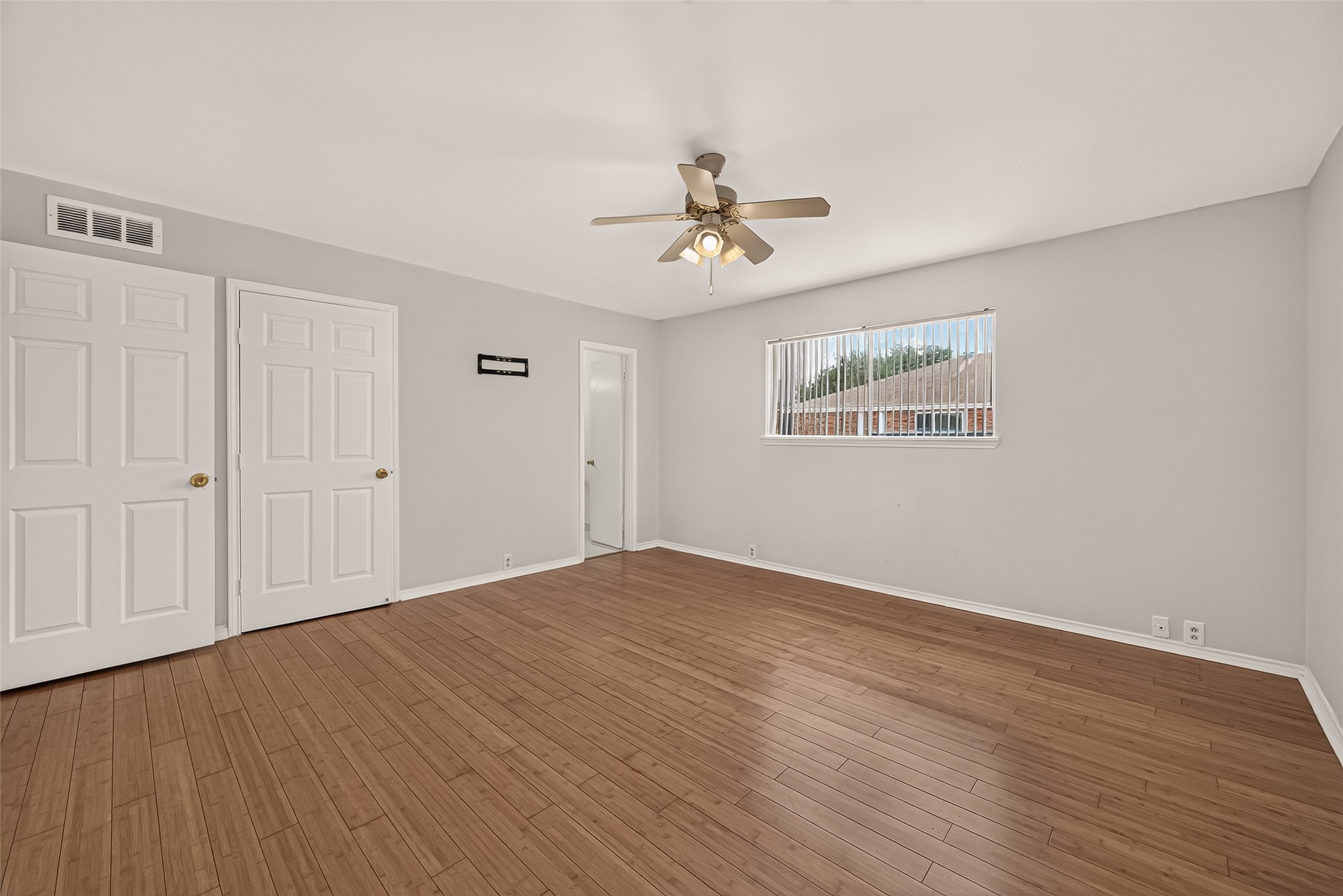 Spacious bedroom with warm wood-style flooring, neutral walls, and ample natural light from the large window, featuring a ceiling fan and dual closets for convenience.