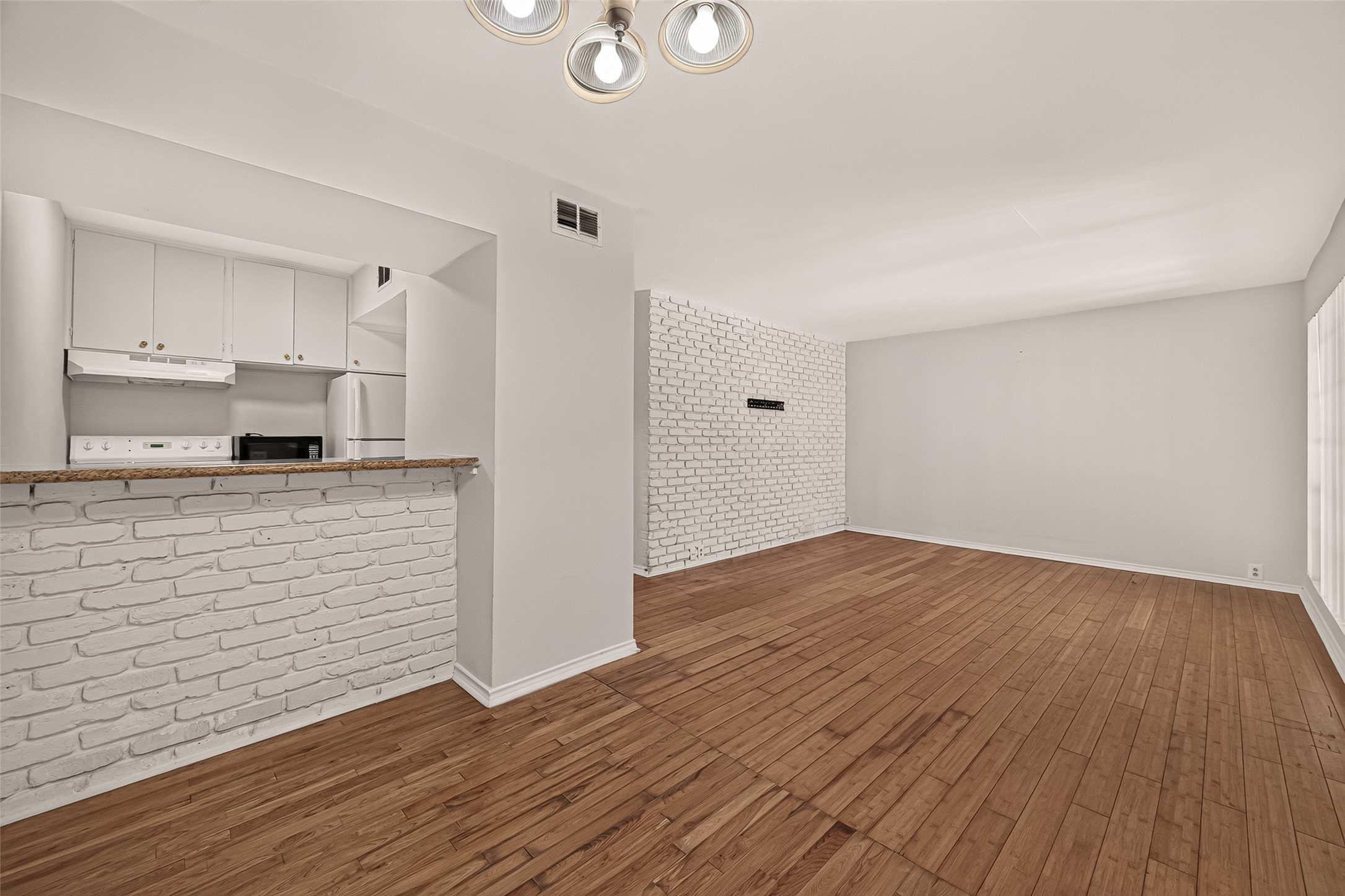 Bright and spacious living area with stylish white brick accents, hardwood floors, and an open kitchen featuring modern appliances and a convenient breakfast bar.