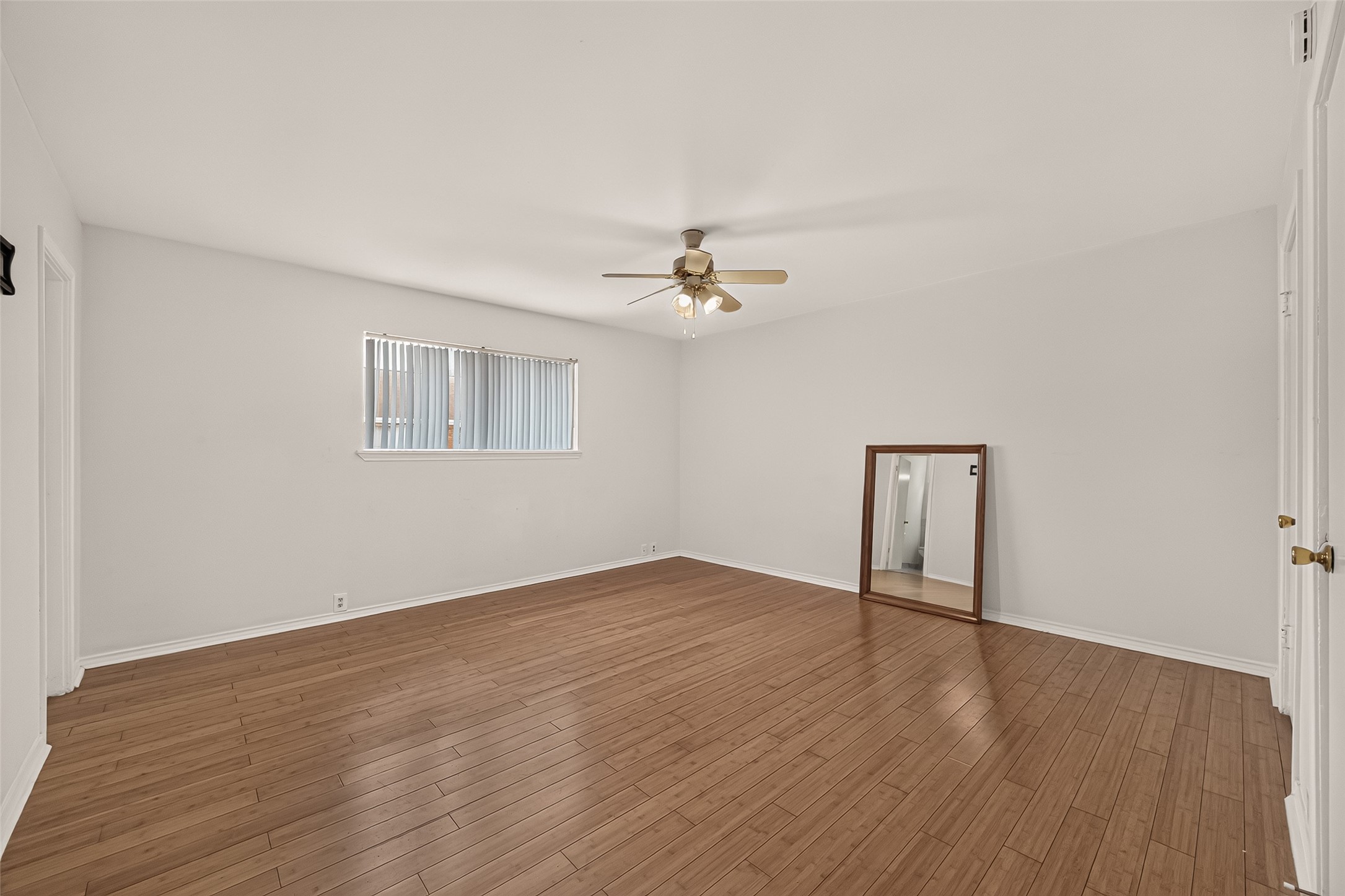 Spacious room with elegant wood flooring, abundant natural light from a large window, and a ceiling fan for comfort.