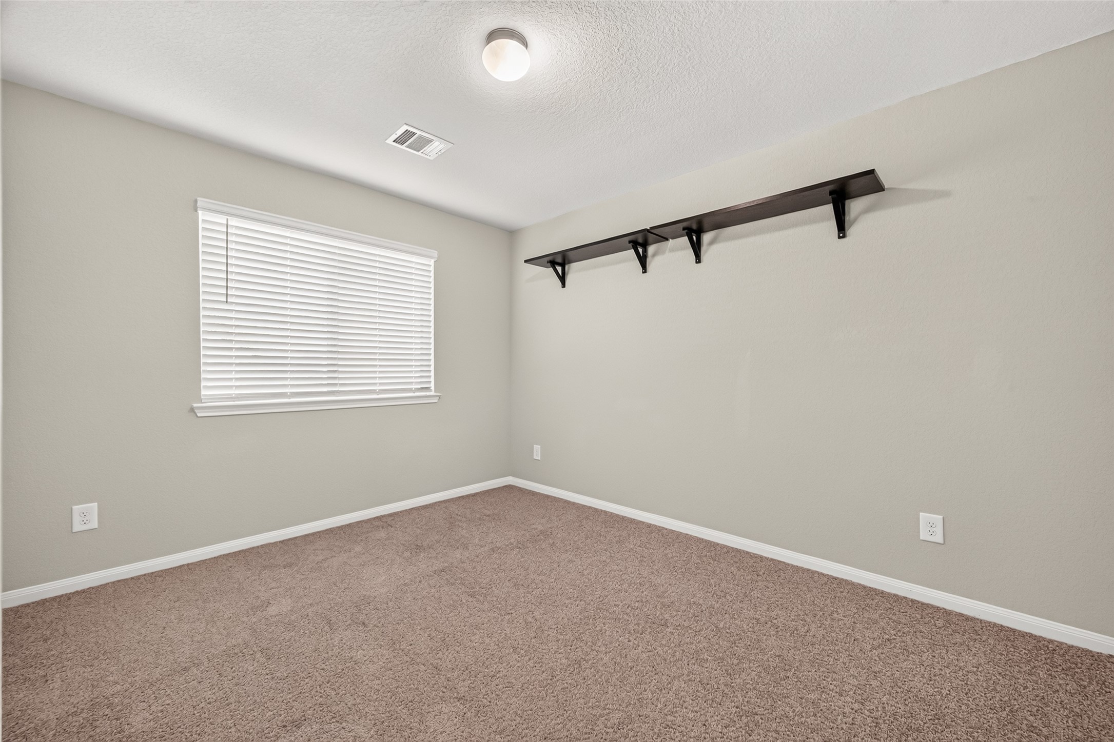 Bedroom  3 with plush carpeting, soft neutral walls, and ample natural light from a large window, featuring a sleek wall-mounted shelf for added storage.