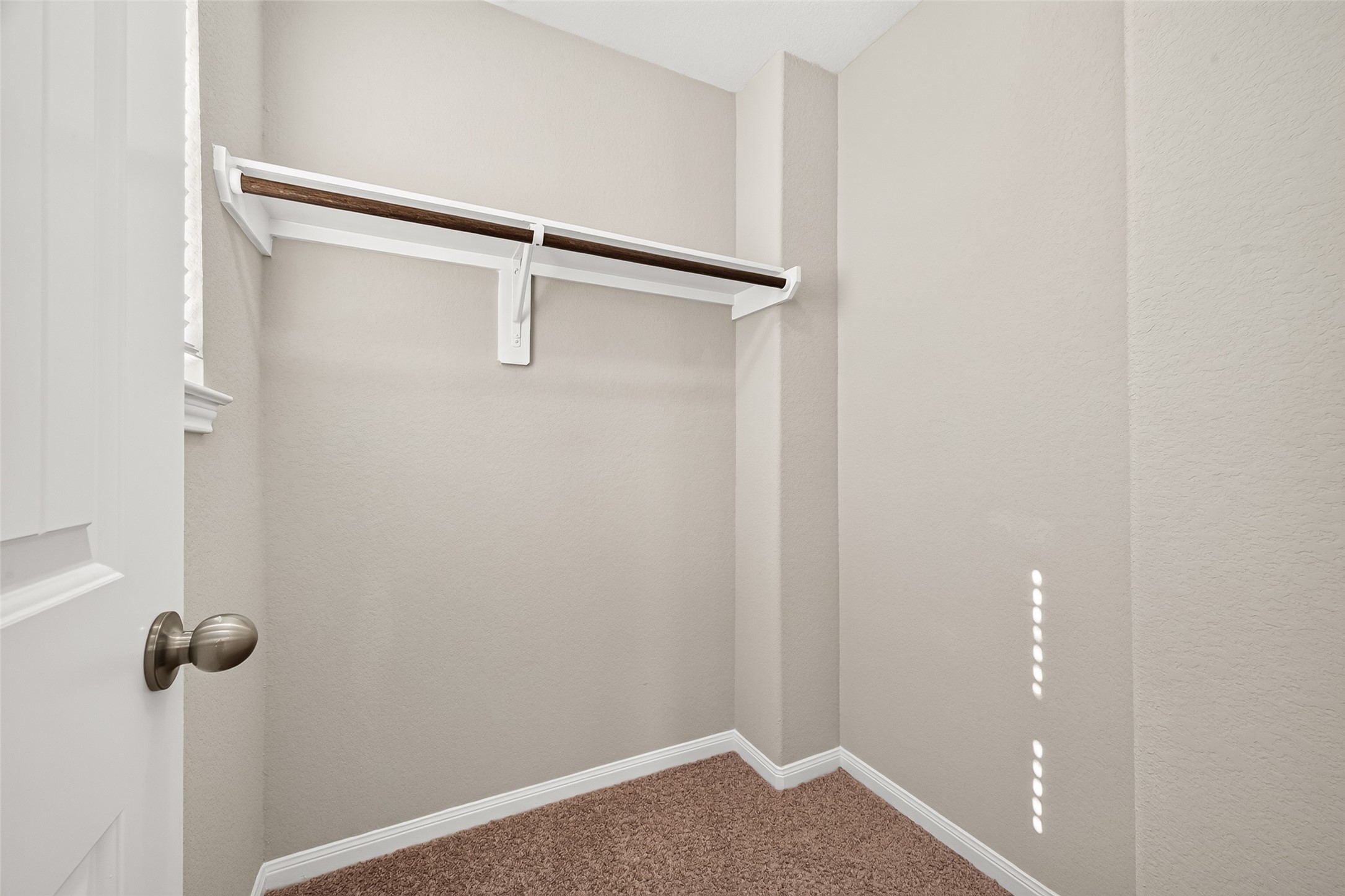 Good size walk-in closet with a sturdy wooden rod for ample hanging storage, complemented by soft carpeting and neutral walls.