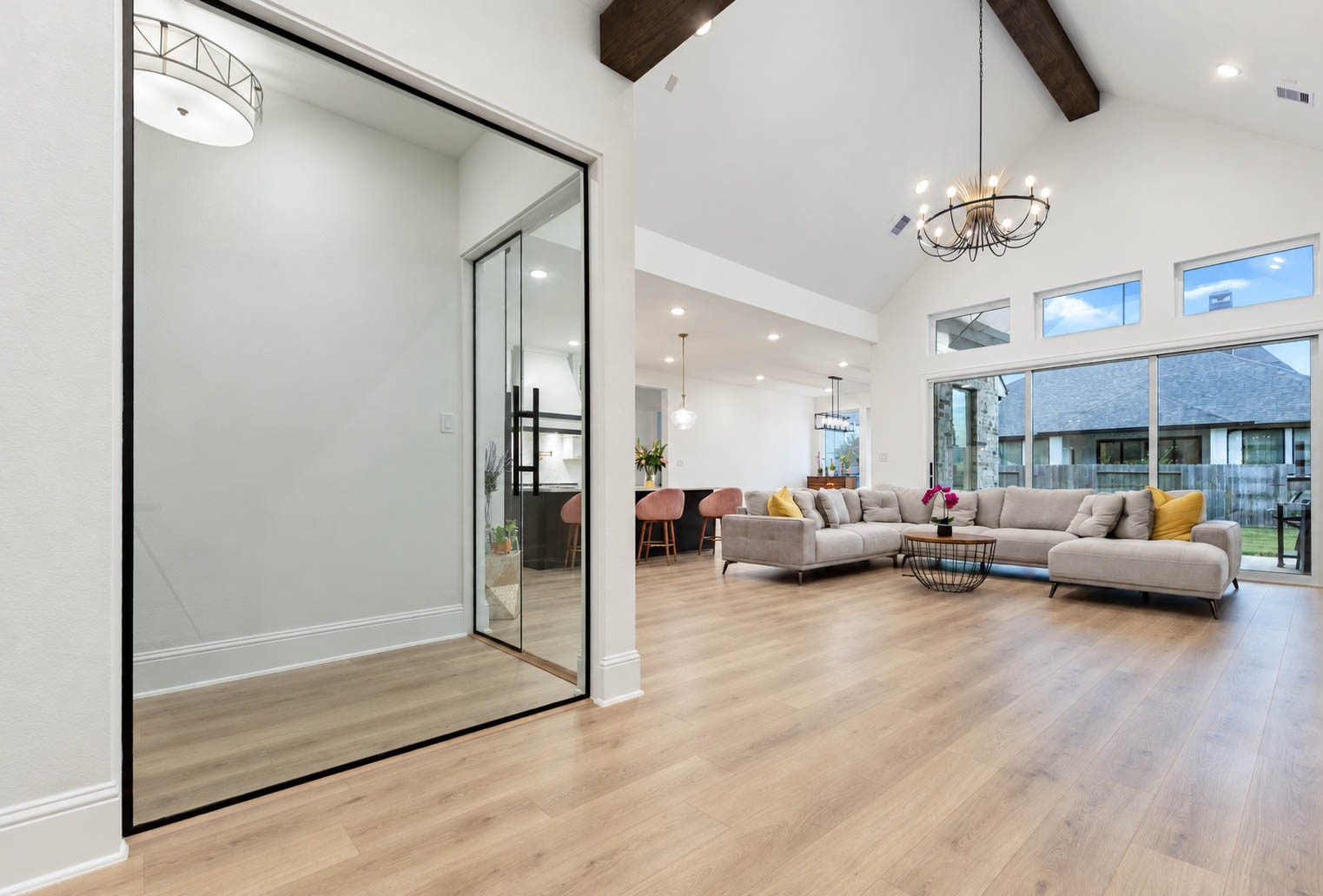 Spacious open-concept living area with vaulted ceilings, showcasing a cozy sectional, elegant chandelier, and abundant natural light from sliding doors overlooking the backyard. Pay attention to the glass enclosed bar! Simply Wow.