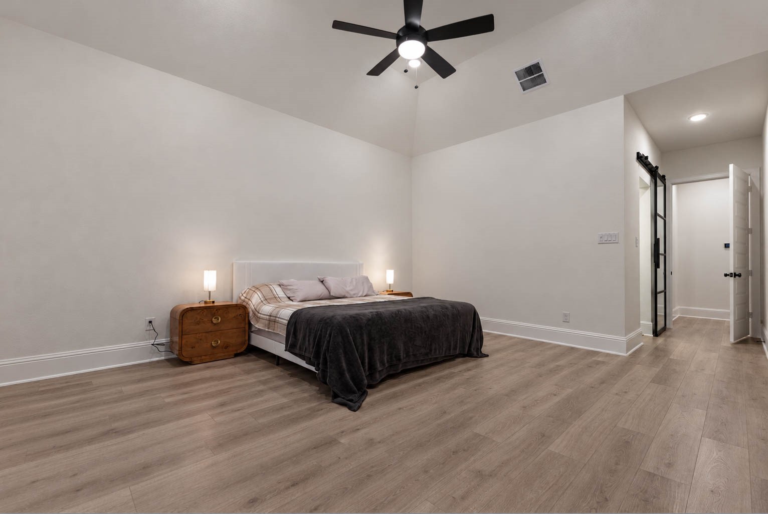 Spacious primary bedroom with high ceilings, featuring elegant wood flooring, a modern ceiling fan, and a sleek barn door leading to an en-suite bathroom.