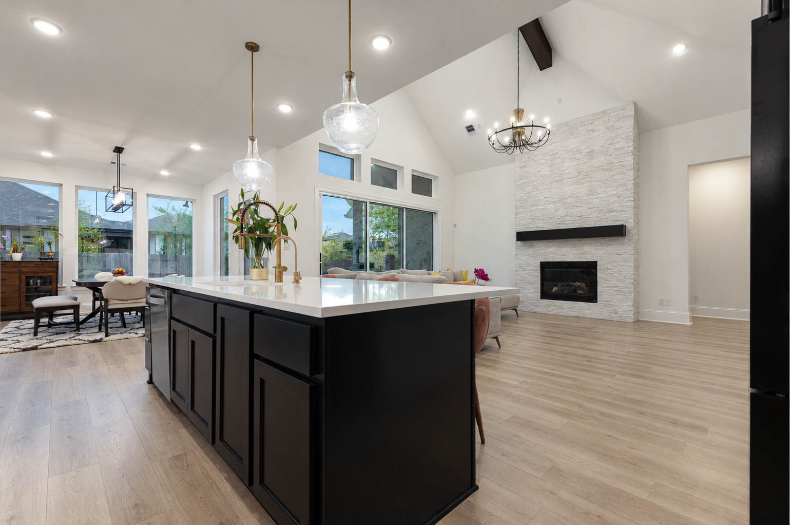 Spacious open-concept living area with high vaulted ceilings, featuring a modern kitchen island, elegant light fixtures, and a cozy stone fireplace. Large windows and sliding doors fill the space with natural light, offering a seamless flow to the dining area. Spacious open-concept living area with high vaulted ceilings, featuring a modern kitchen island, elegant light fixtures, and a cozy stone fireplace. Large windows and sliding doors fill the space with natural light, offering a seamless flow to the dining area.