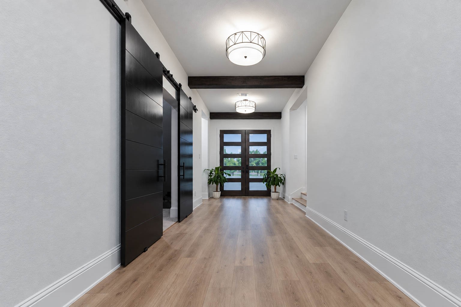 Stunning entryway with elegant wood flooring, modern lighting, and sleek black barn doors for the media room, leading to a bright space with large windows and natural light.