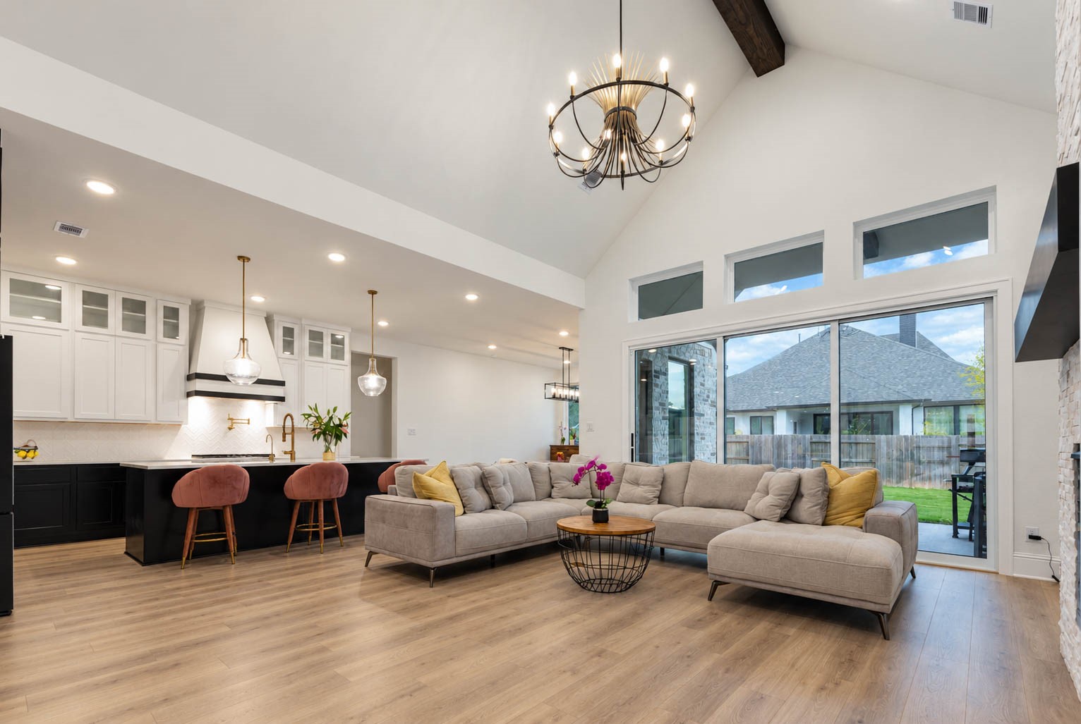 Spacious open-concept living area with vaulted ceilings, elegant chandelier, and modern kitchen featuring a stylish island with seating and pendant lights. Expansive windows enhance natural light and offer a view of the backyard. Sliding doors.