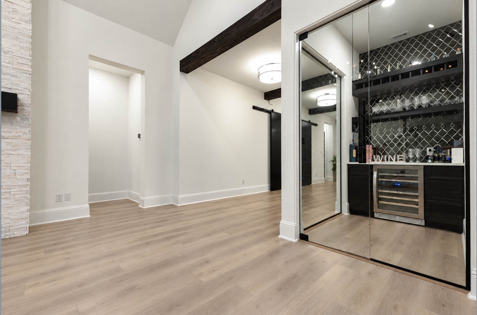 Elegant room featuring a sleek wine bar with mirrored sliding doors, chic backsplash, and modern lighting, complemented by warm wood flooring and exposed beams. Cheers!