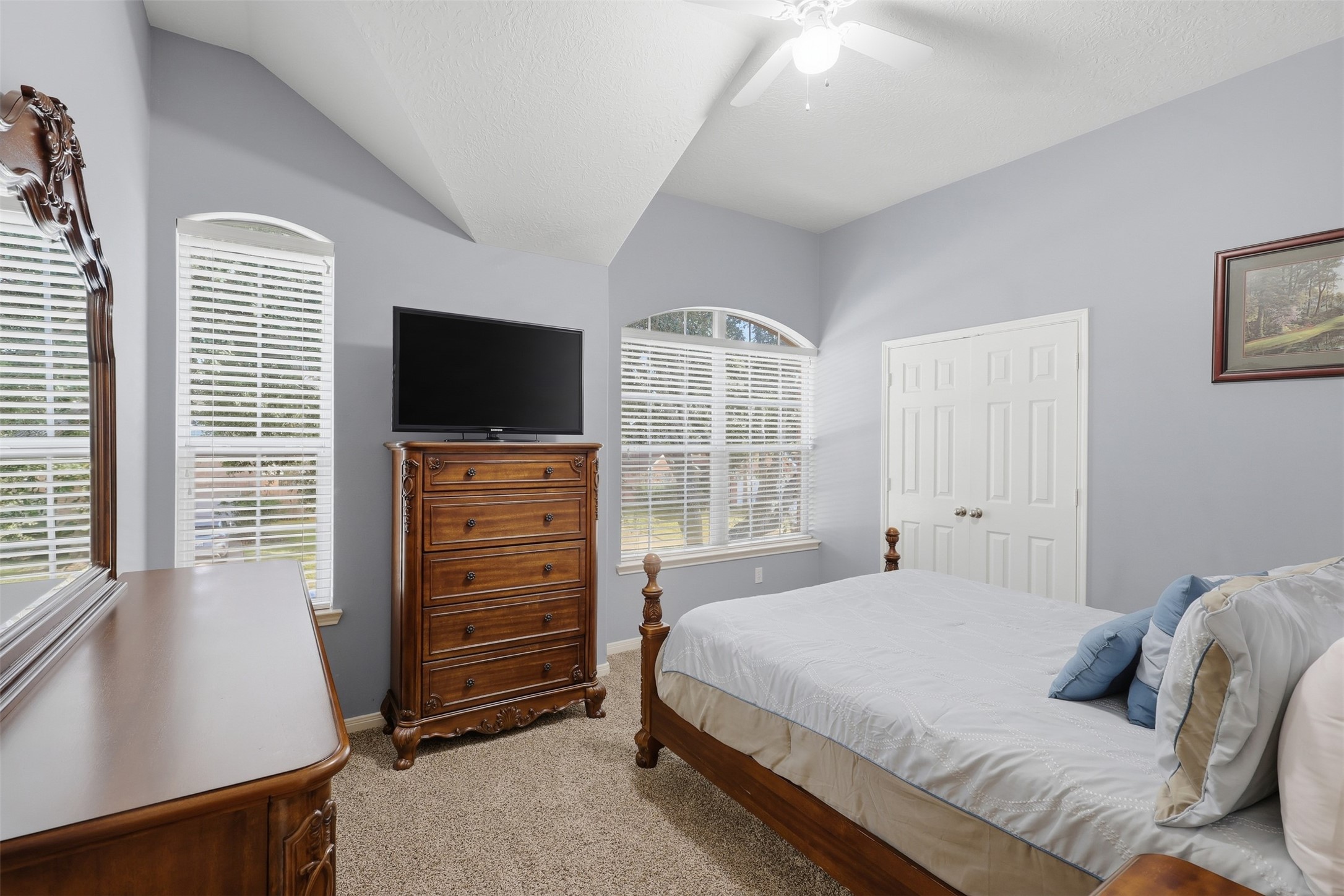 Spacious secondary bedroom with plush carpeting, multiple windows adorned with plantation-style blinds, a ceiling fan, and ample floor space. The bright, inviting layout comfortably accommodates a bedroom set, dresser, and personal touches. French doors open to a large, well-designed walk-in closet.