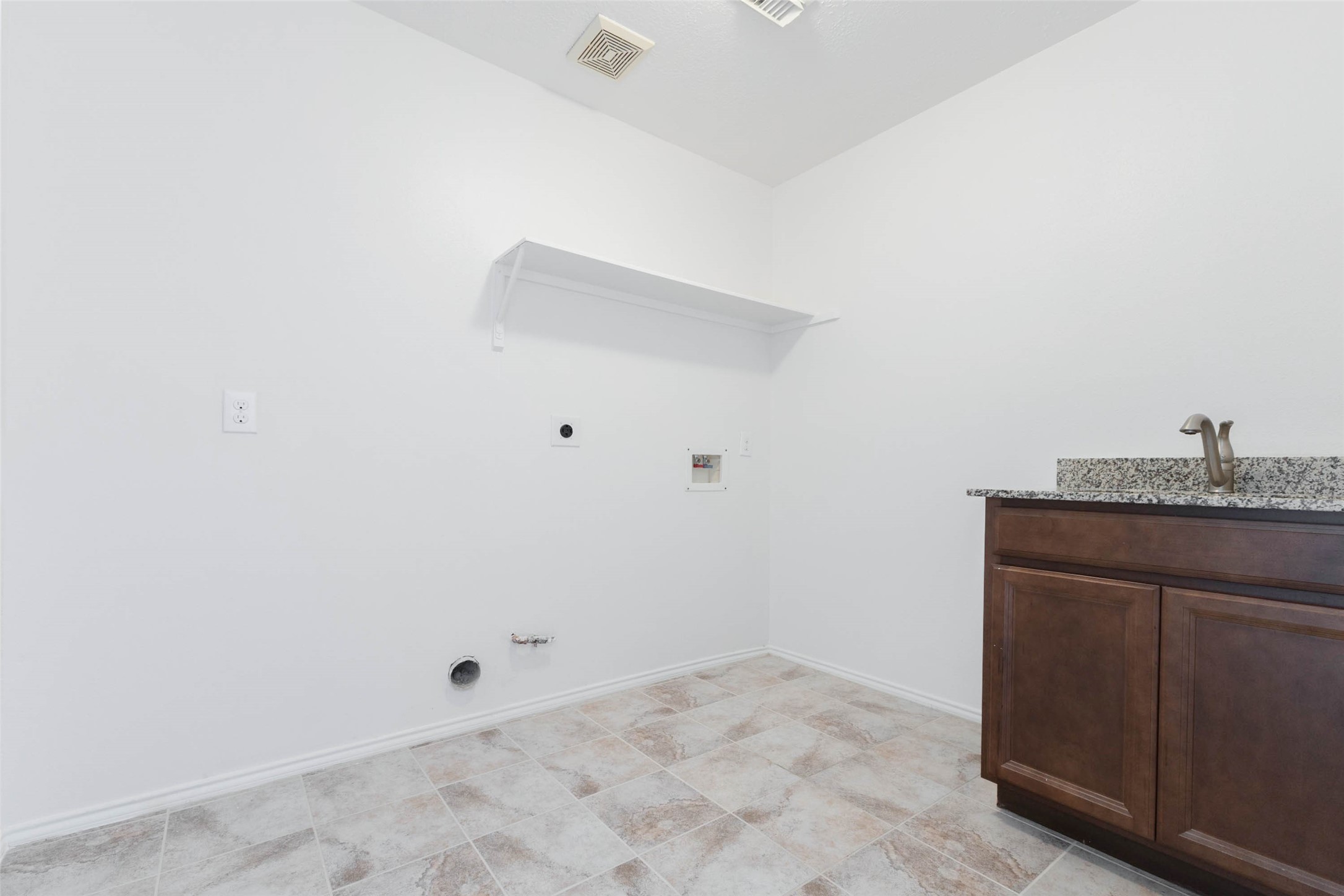 This spacious laundry room offers a convenient & functional workspace featuring a granite-topped utility sink with rich wood cabinetry for additional storage. Neutral tile flooring, fresh interior paint, & overhead shelving complement the practical design. *