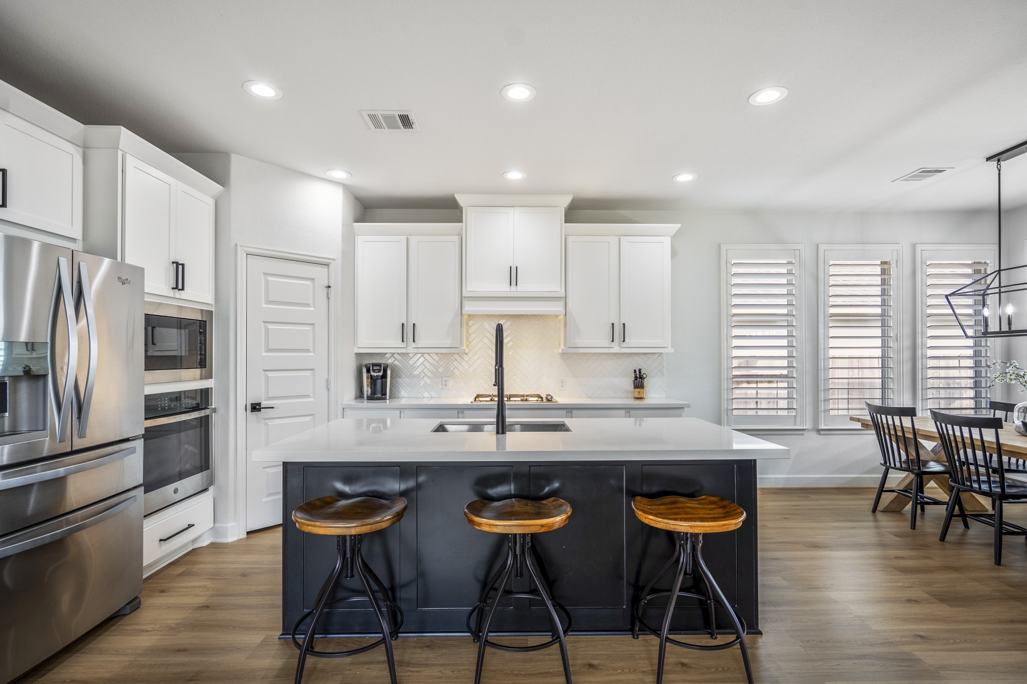 This modern kitchen features sleek stainless steel appliances, white cabinetry, and a spacious island with seating. The open layout includes a dining area with large windows, providing ample natural light and a cozy, inviting atmosphere.