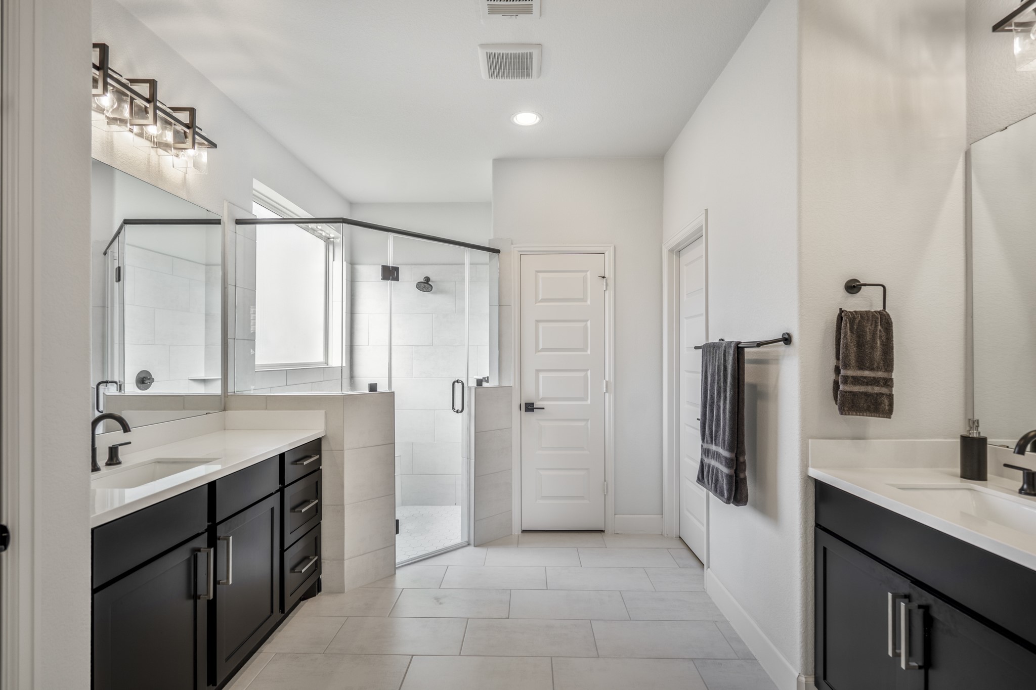 This modern primary bathroom features dual vanities with sleek black cabinets, a spacious glass-enclosed shower, and elegant lighting fixtures, all set against crisp white walls and tile flooring.