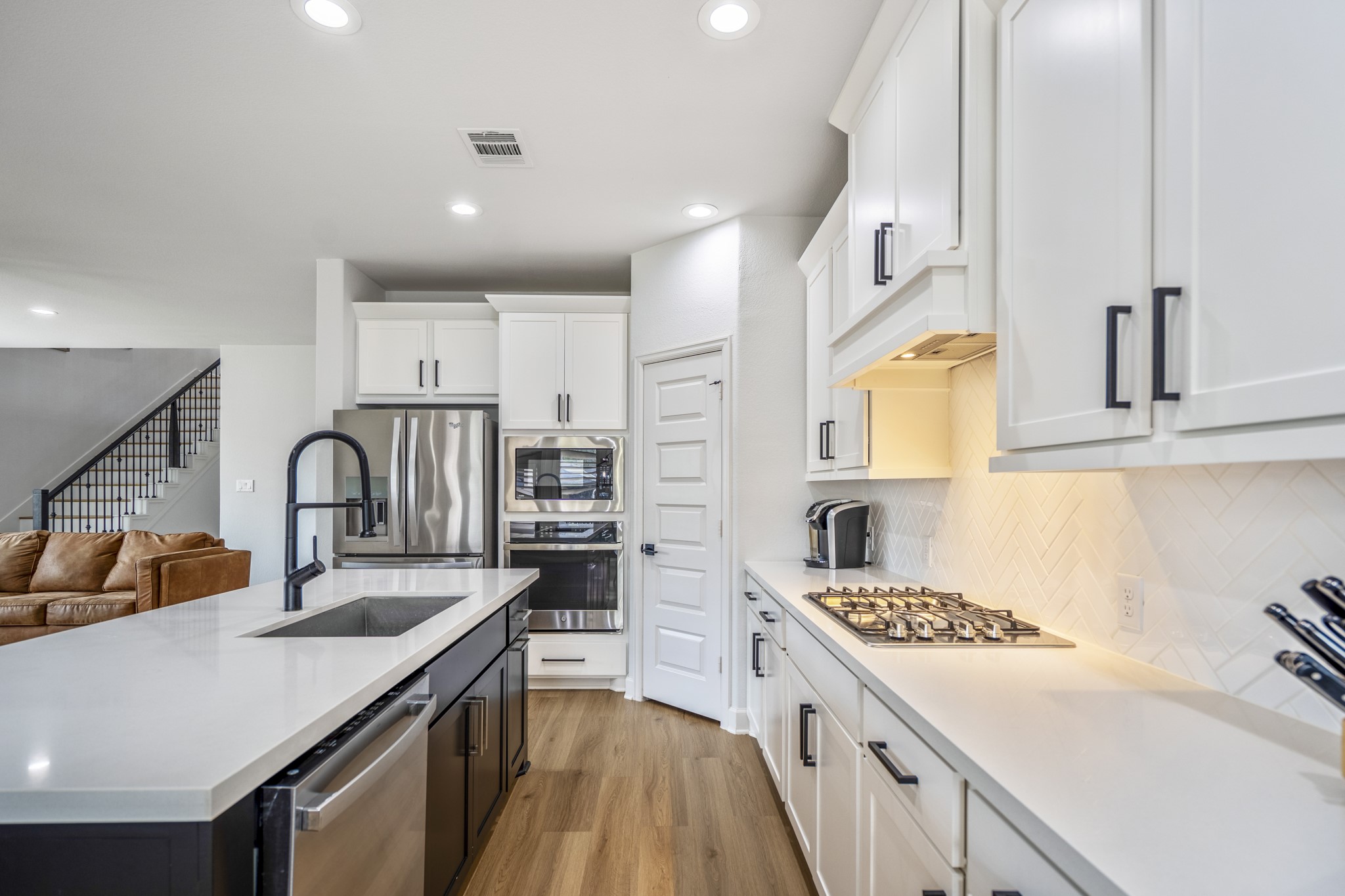 It features a gas stove, built-in oven, and ample counter space, all illuminated by recessed lighting. The open layout flows into the living area with a glimpse of a staircase.