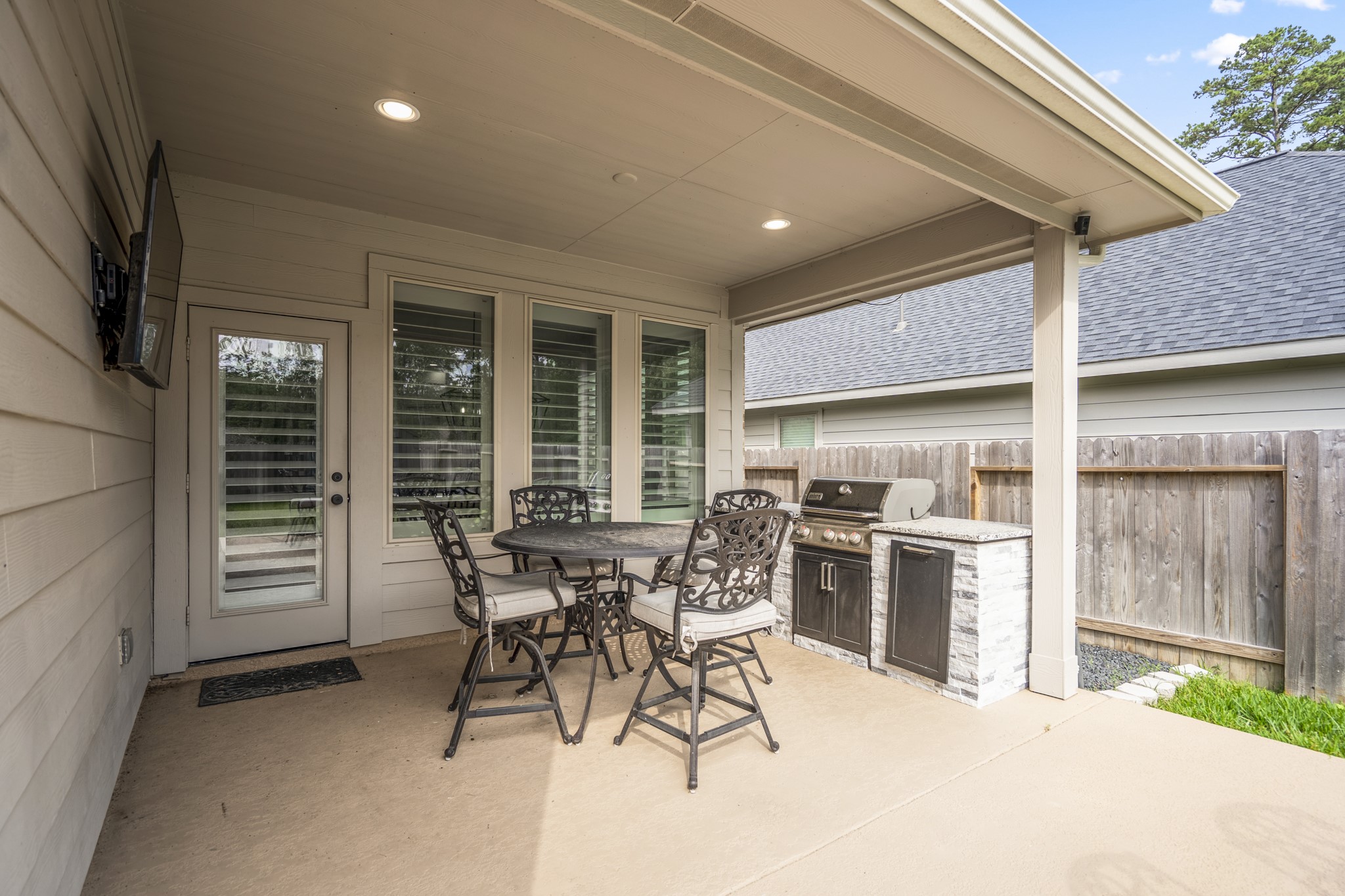 This photo showcases a cozy covered patio with a built-in grill, outdoor seating, and a mounted TV, perfect for entertaining or relaxing.