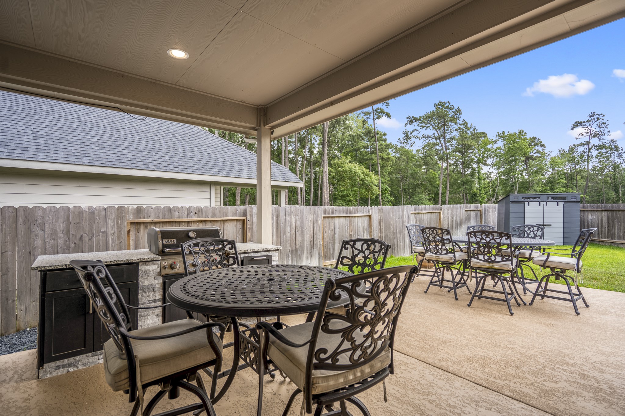The photo features a spacious covered patio with a built-in grill and overlooks a fenced backyard with a storage shed, offering privacy and a lush tree-lined view.