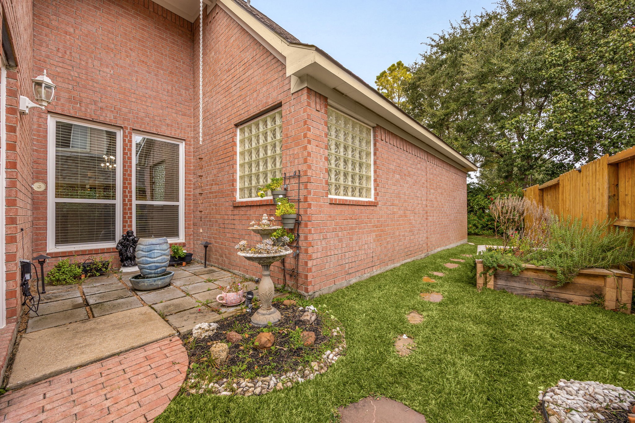 This beautifully landscaped corner showcases a stepping-stone path leading to a built-in stone planter bed, perfect for a home garden. Surrounded by classic four-sided brick architecture and private glass-block windows, it offers another peaceful outdoor pocket to enjoy your serene surroundings.