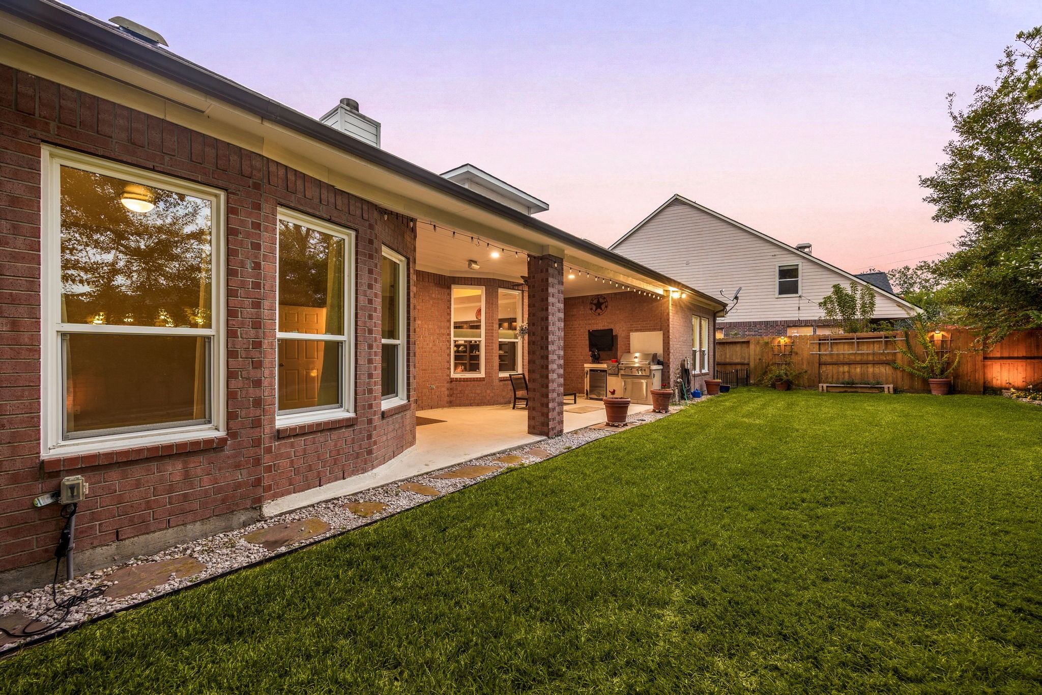 This view showcases the impressive length of the covered outdoor living space, framed by a sprawling, beautifully manicured green lawn. The combination of classic full-brick elevation, glowing interior windows, and a peaceful evening sky perfectly highlights the exceptional size and premium outdoor lifestyle this Cinco Ranch property provides.