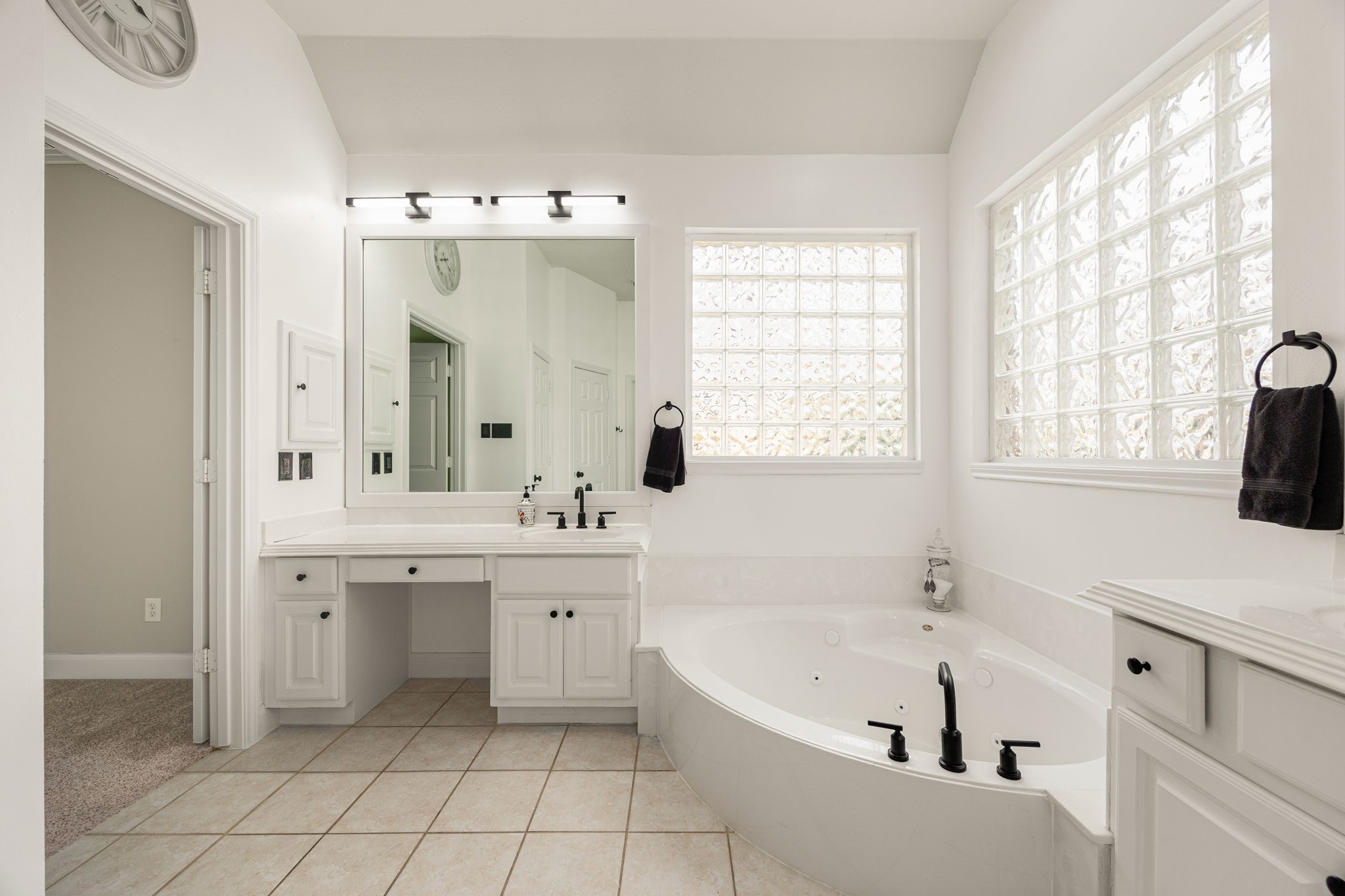 A dedicated vanity area with a knee-space makeup counter sits adjacent to the luxurious soaking tub, while premium glass-block windows flood the room with natural light without compromising privacy. The doorway opens directly into the massive primary closet.