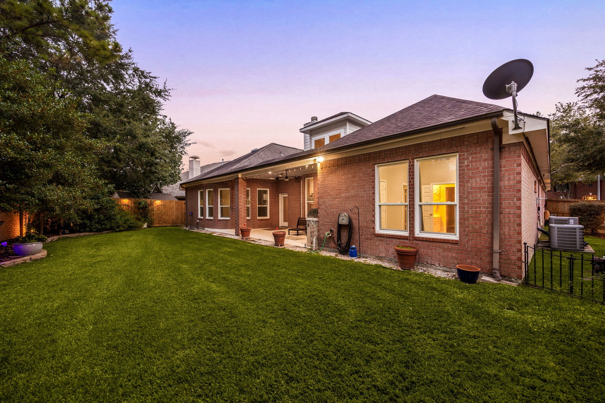This view showcases the impressive length of the covered outdoor living space, framed by a sprawling, beautifully manicured green lawn. The combination of classic full-brick elevation, glowing interior windows, and a peaceful evening sky perfectly highlights the exceptional size and premium outdoor lifestyle this Cinco Ranch property provides.