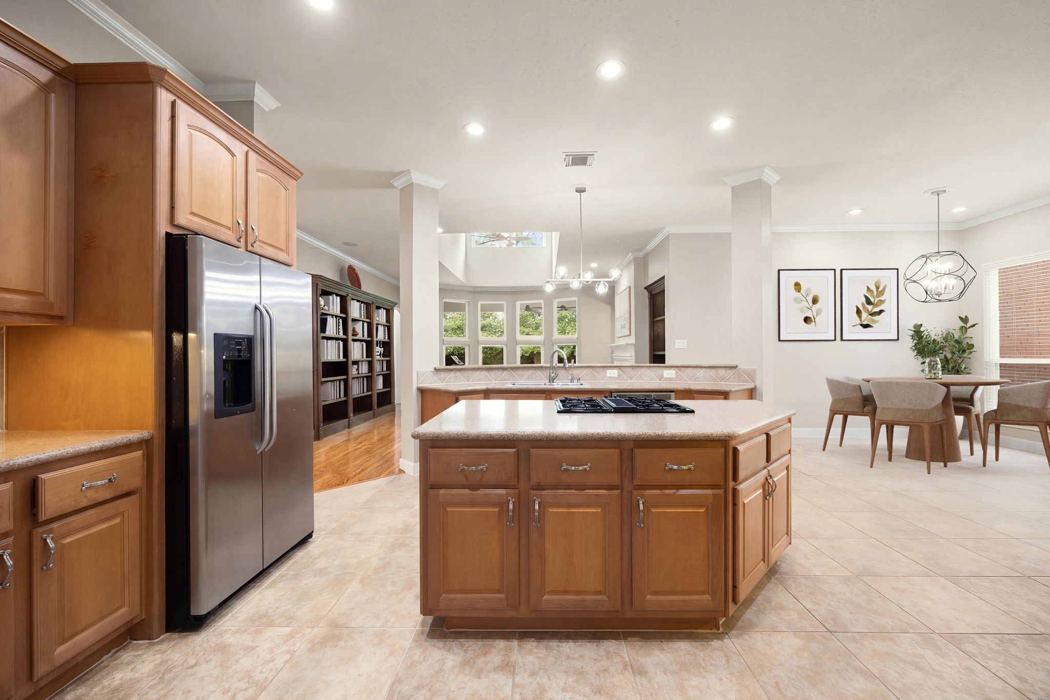 This expansive kitchen workspace features an oversized center island with a built-in cooktop, premium stainless steel appliances, and a matching refrigerator. The sprawling layout seamlessly connects the kitchen to the casual dining area, formal dining space, and main living room, offering the perfect arrangement for daily meals and grand-scale holiday hosting.