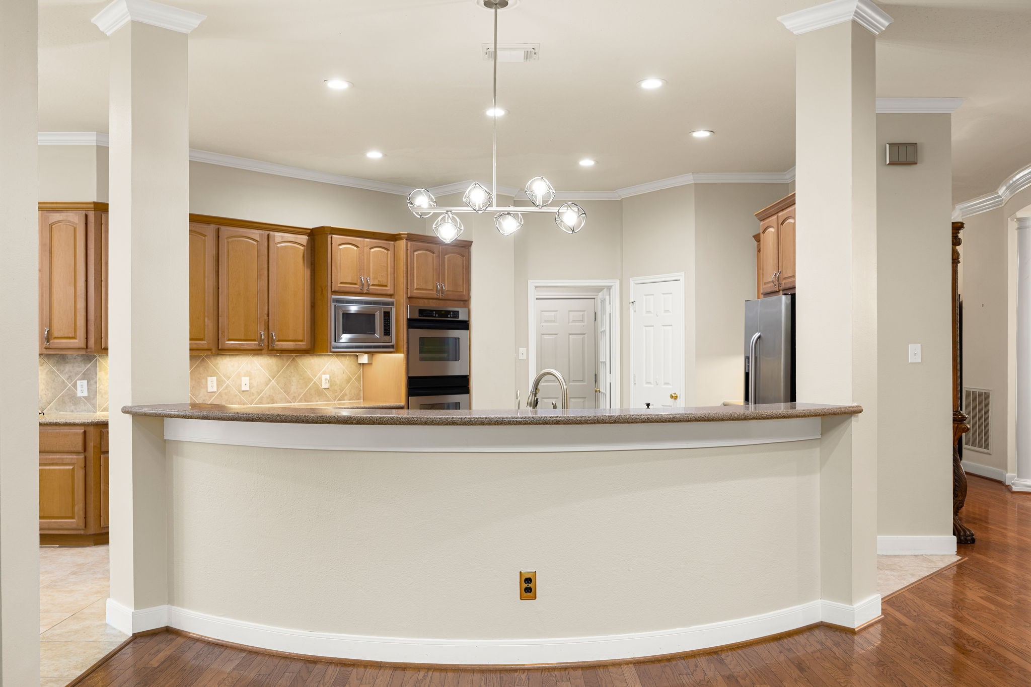 This stunning gourmet kitchen features rich wood cabinetry, built-in stainless steel appliances, and premium recessed lighting. The dramatic, curved breakfast bar overlooks the living spaces, anchored by an elegant, modern branching chandelier that adds a sophisticated touch to the home's open-concept design.
