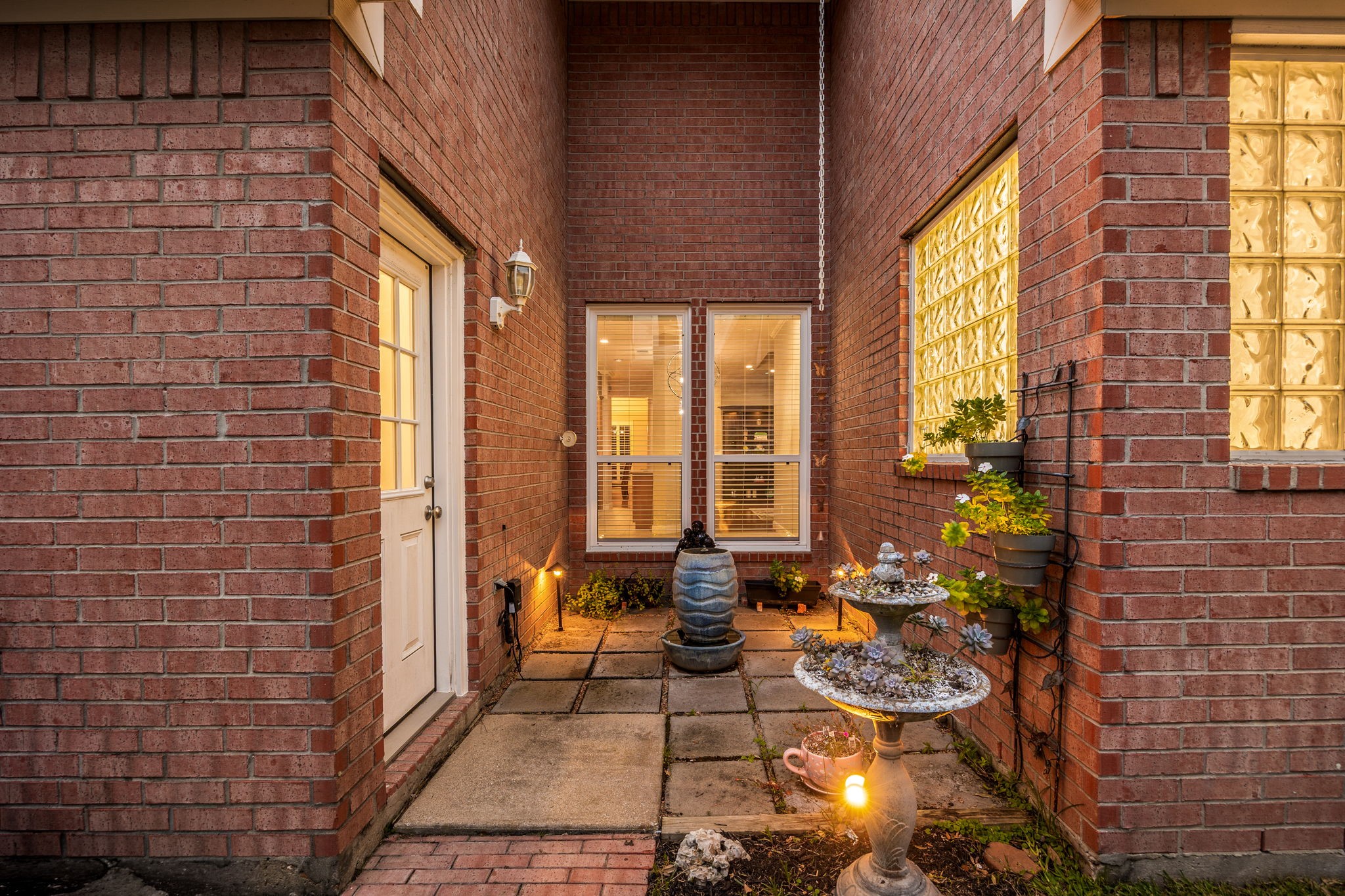 This hidden architectural gem offers an intimate outdoor escape, beautifully laid with stone pavers and glowing under warm exterior lighting. Accessible via a glass-paneled door and overlooked by elegant privacy-glass block windows, this tucked-away courtyard is the perfect quiet nook for a morning coffee or a cozy bistro seating arrangement.