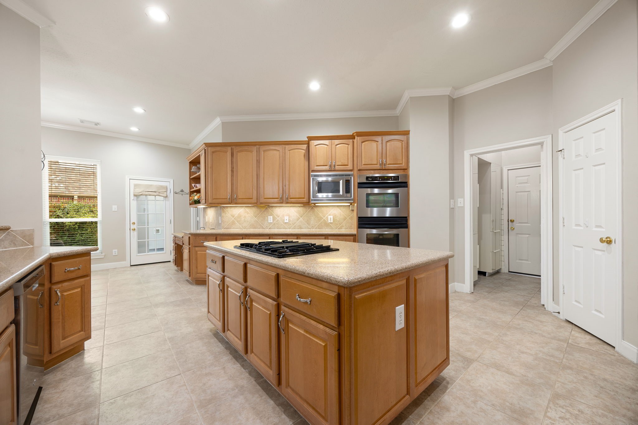 This viewpoint emphasizes the premium built-in stainless steel double ovens and matching microwave seamlessly integrated into the custom cabinetry. Bright recessed lighting combined with under-cabinet task lighting illuminates the polished solid-surface countertops, while a convenient backyard exit door keeps outdoor grilling within arm's reach.