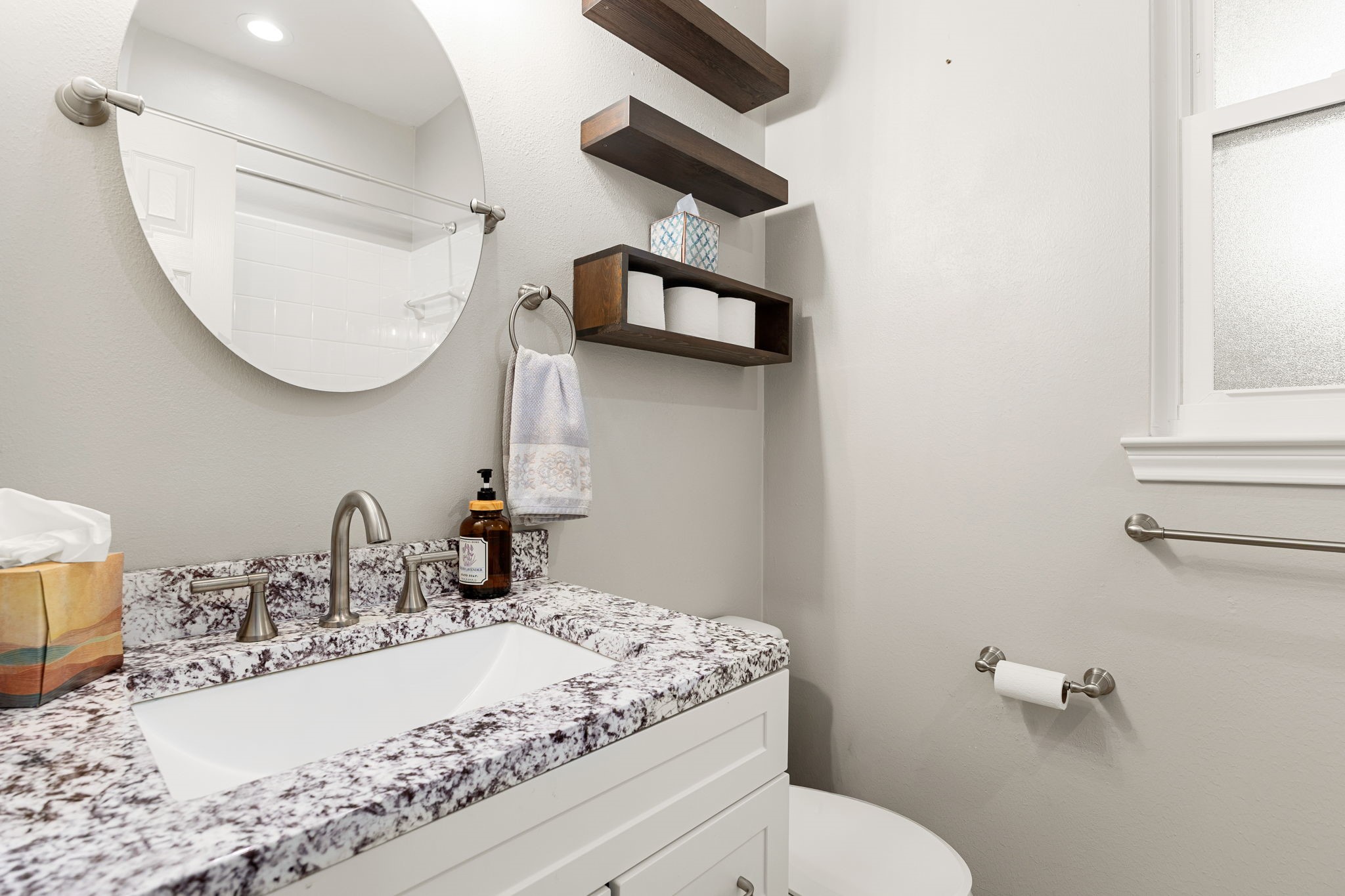 This bath features a premium granite vanity top with striking character, a sleek white cabinet base, and a contemporary frameless circular mirror. This beautifully updated space offers a chic and welcoming environment for family and guests alike.