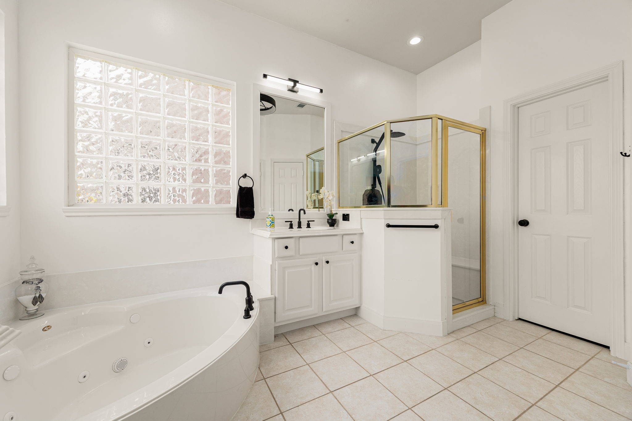 The primary bathroom is a serene oasis featuring a deep, separate jetted soaking tub nestled beneath a bright privacy-glass block window. Complete with a spacious walk-in shower, bright white vanity cabinetry, elegant modern fixtures, and crisp tile flooring, it offers the perfect space to unwind and recharge.
