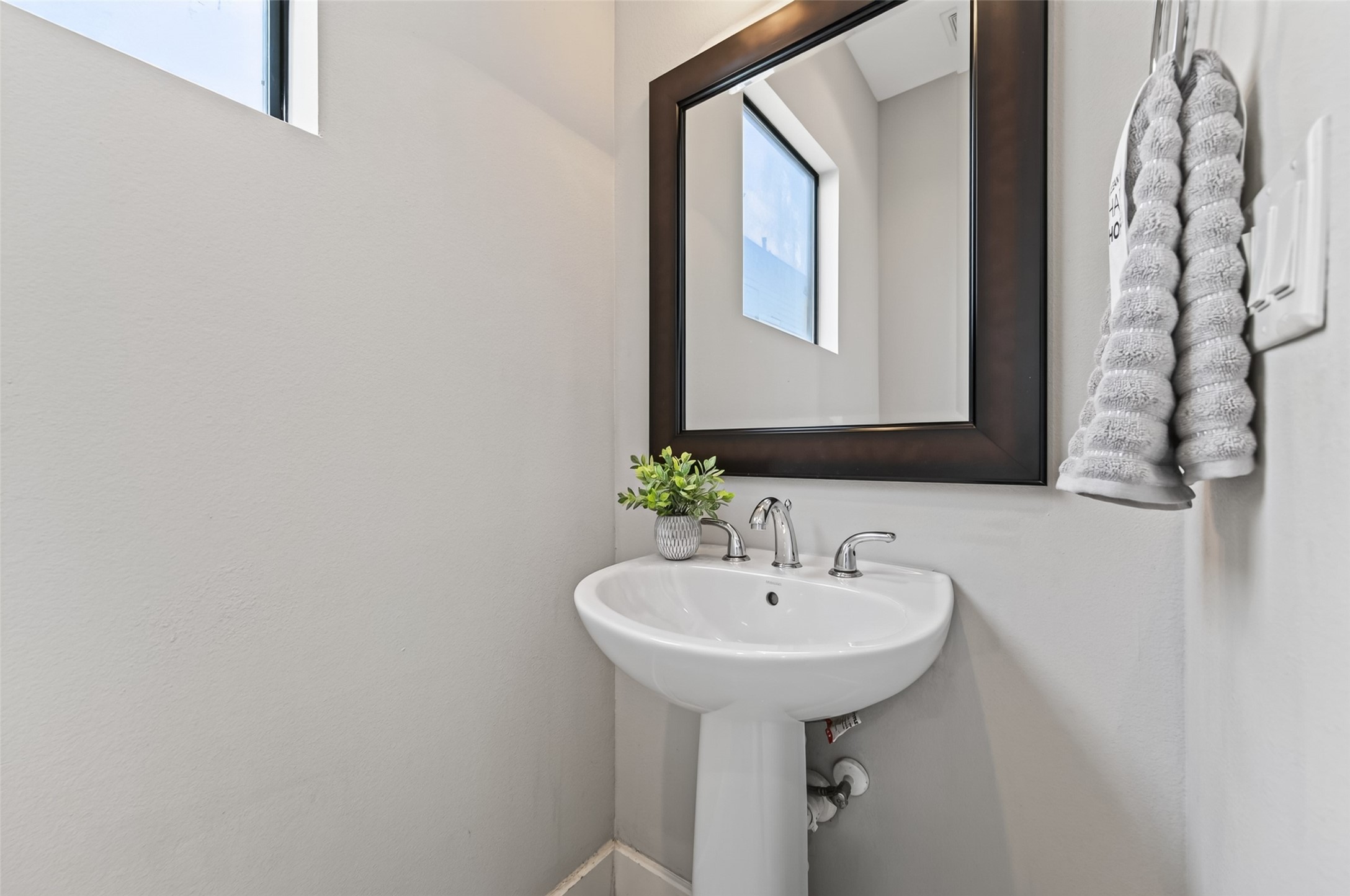 A chic powder room featuring a sleek pedestal sink, modern fixtures, and a large framed mirror, complemented by a bright window and elegant towel display.