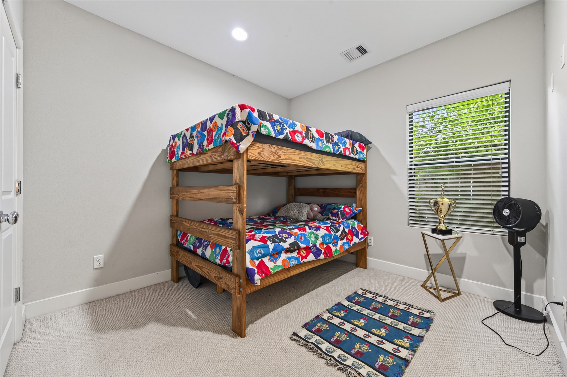 Charming versatile bedroom featuring a cozy wooden bunk bed, colorful decor, and a playful rug. The room is brightened by natural light from a window, complemented by a small play table and storage units.