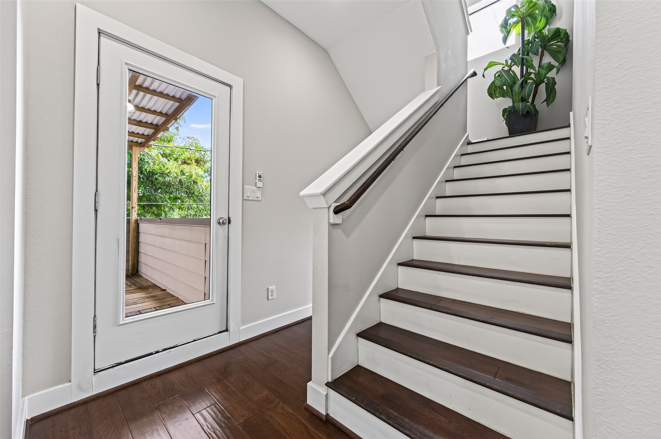Bright entryway featuring elegant dark wood flooring, a sleek staircase with white accents, and a glass door leading to a cozy outdoor deck.