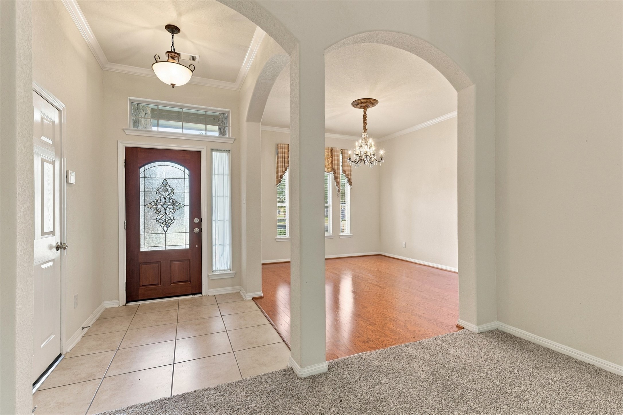Elegant entryway with a stunning wooden door featuring decorative glass, leading to a bright dining area with hardwood floors and a classic chandelier.