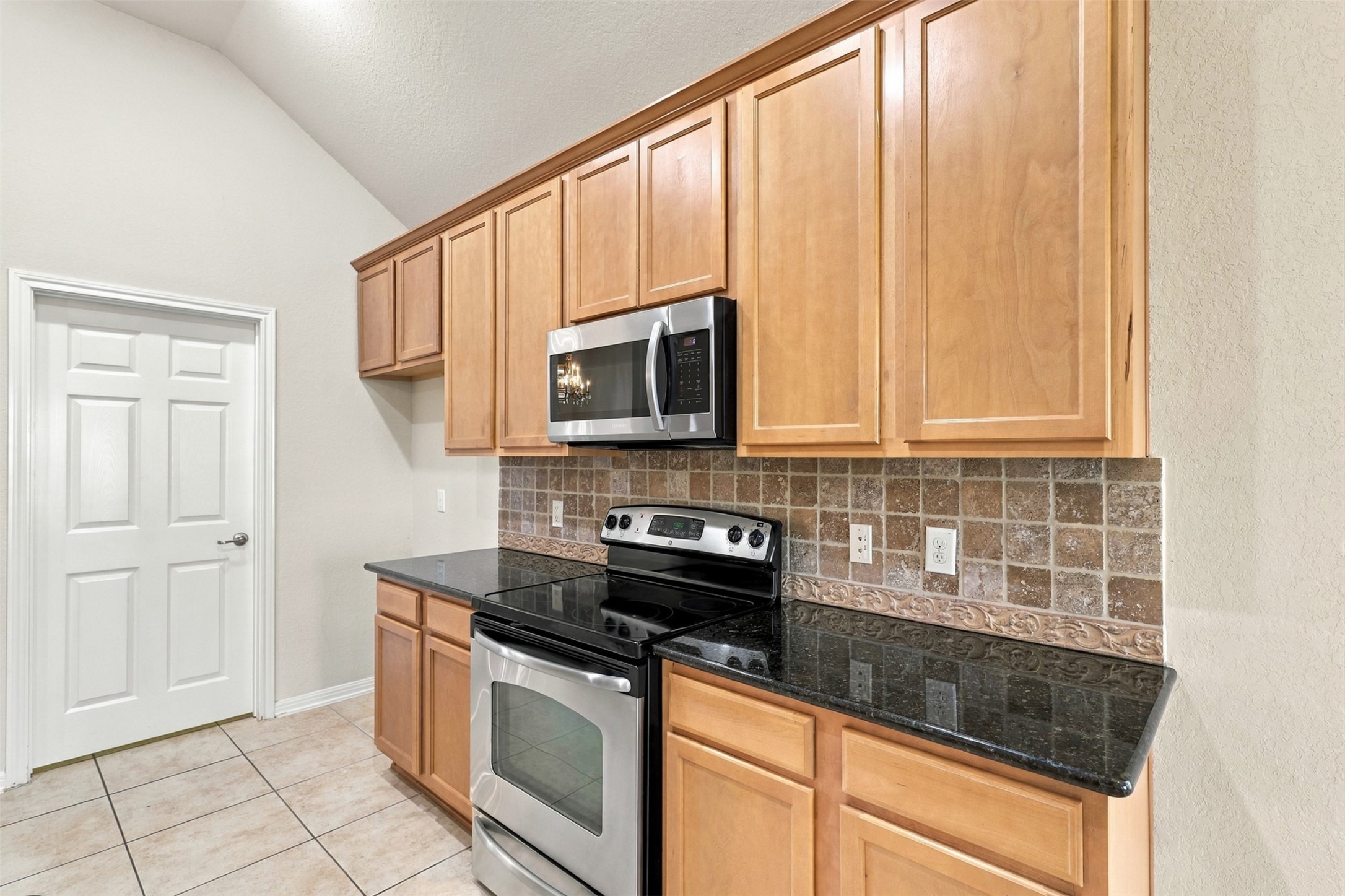 Another view of the kitchen with sleek black granite countertops, stainless steel appliances, and ample light wood cabinetry for storage.
