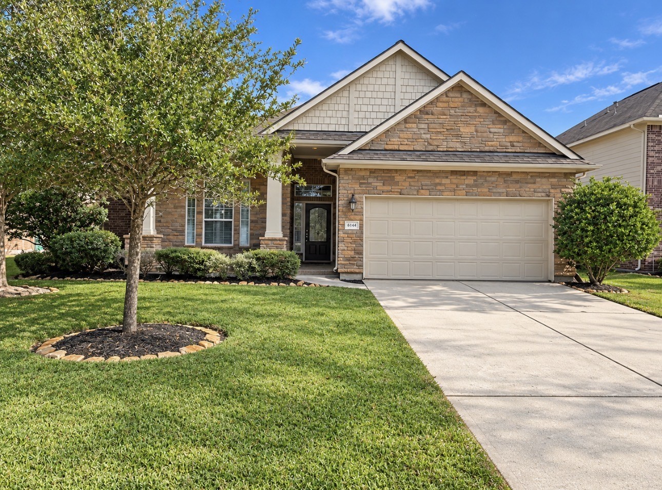 The perfect single-story home with a stone facade and inviting front porch. Features a well-maintained lawn, mature tree, and a spacious two-car garage.