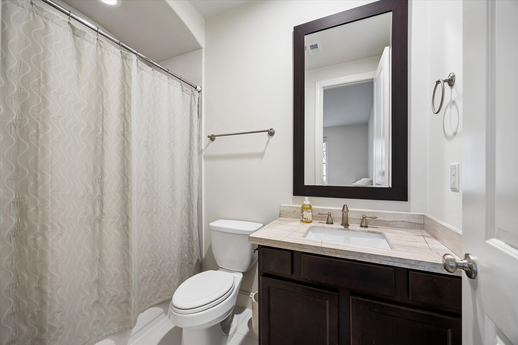 En suite bath for the first floor bedroom provides added privacy and convenience, complete with a tub and shower combination, stone topped vanity, and dark wood cabinetry.