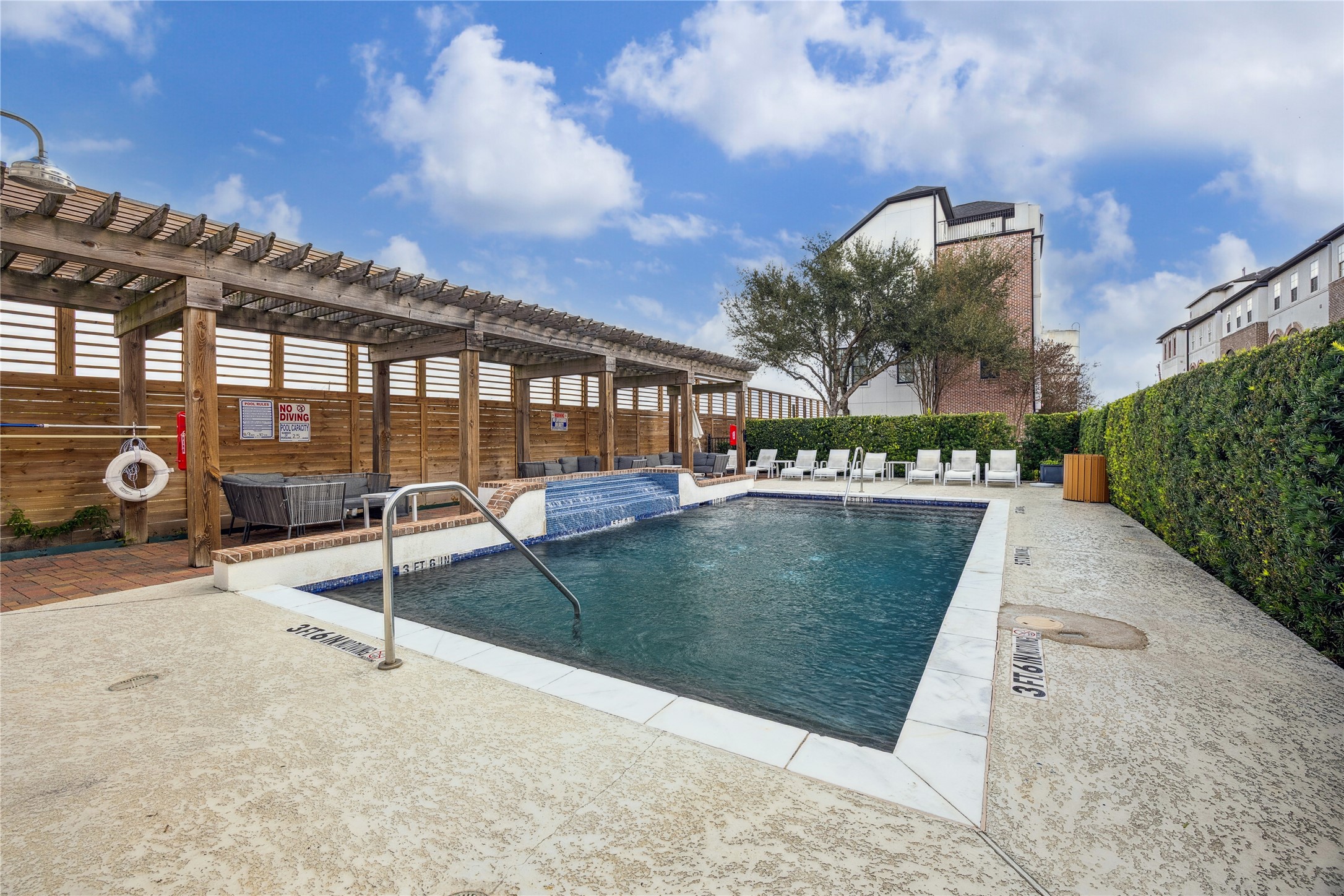 A resort style pool is framed by a wood pergola, brick accents, and cushioned lounge seating for shaded relaxation. Open deck space and privacy hedges create a clean, inviting setting for warm weather afternoons.