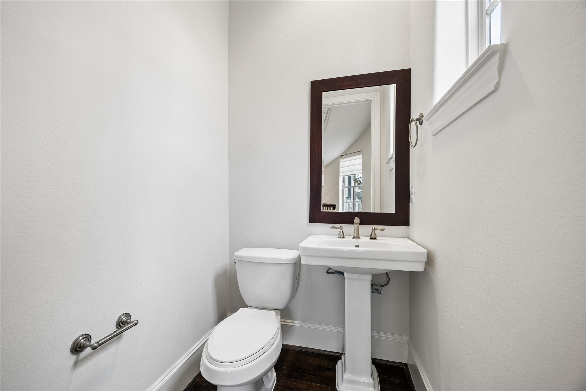 Natural light from a high window brightens the powder bath, finished with a classic pedestal sink, framed mirror, and hardwood flooring.
