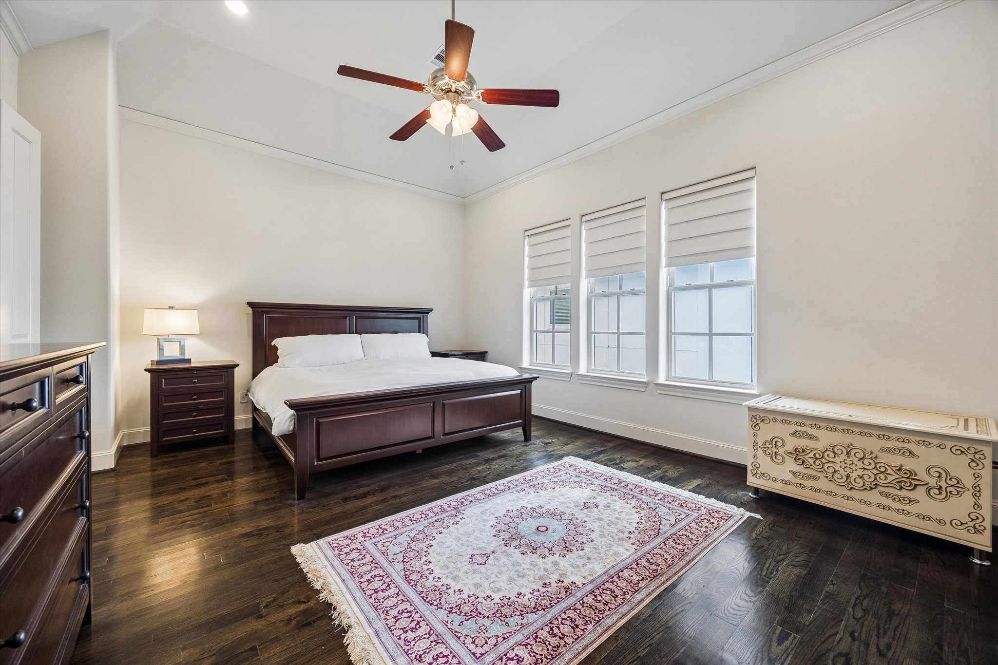 Three tall windows bring natural light into the spacious primary suite, complemented by rich hardwood floors, a tray ceiling, and generous wall space.
