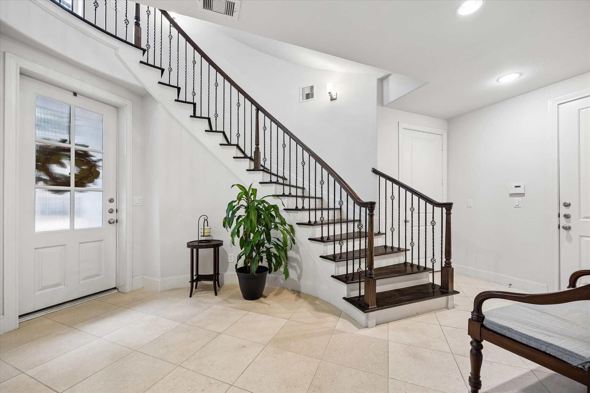 A graceful staircase with dark wood treads and wrought iron detailing creates an architectural focal point upon entry. Light stone floors and bright white walls keep the foyer feeling open and polished.