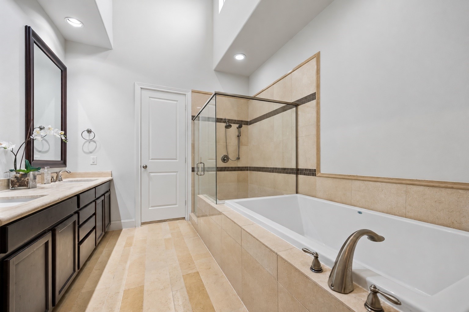 A spacious primary bath pairs warm stone tile with dark cabinetry and a dramatic vaulted ceiling. An oversized soaking tub and separate glass enclosed shower create a comfortable retreat, with direct access to one of the two primary closets.