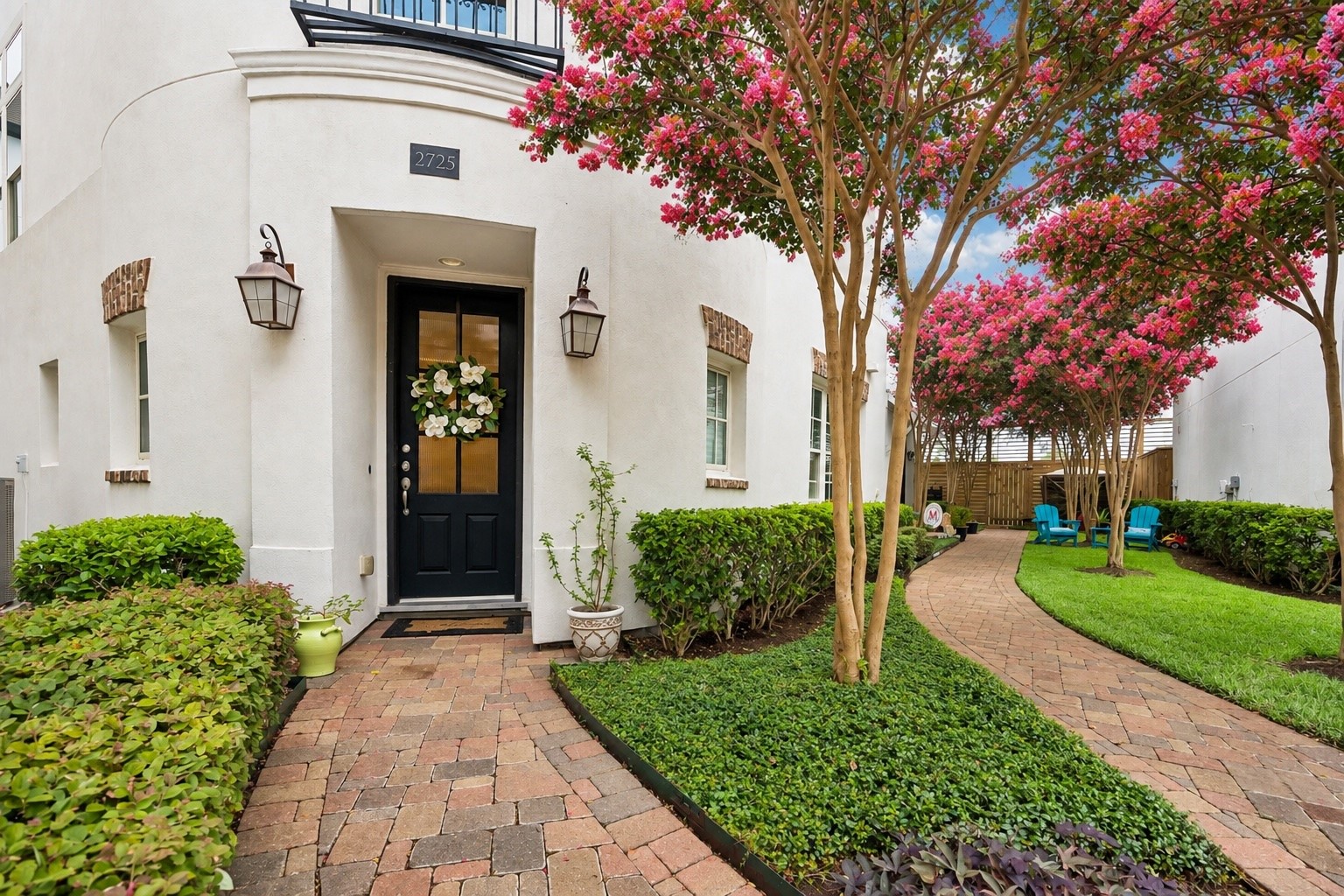 A charming brick walkway winds through manicured landscaping beneath vibrant crepe myrtles, creating a private, garden like approach to the home. The recessed entry and classic lanterns add warmth to the crisp stucco exterior.
